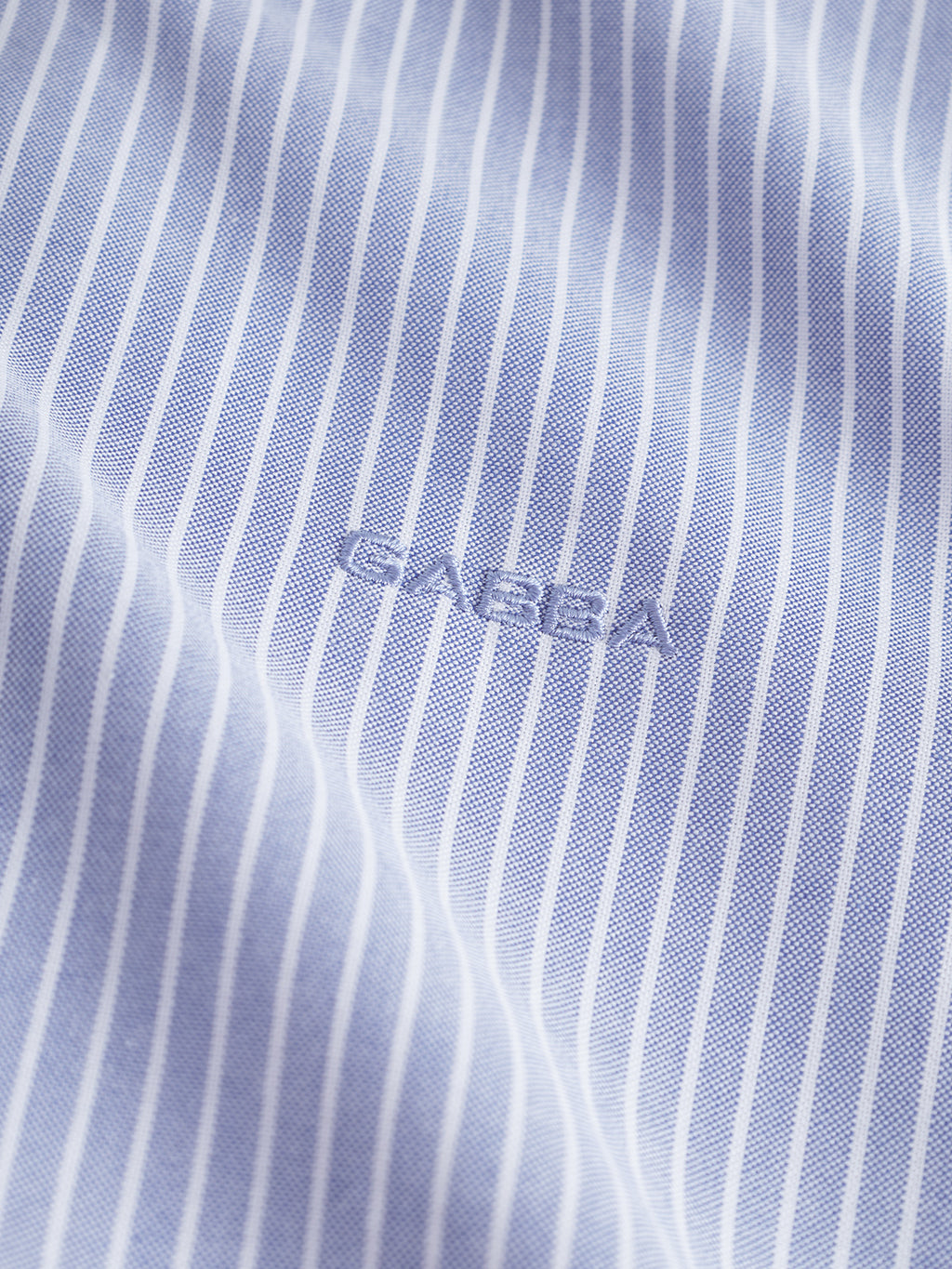 Close-up of a blue and white pinstripe fabric from the Gabba Sven Reg Crest LS - Blue Stripe shirt, featuring Gabba embroidered in matching blue thread at the center.