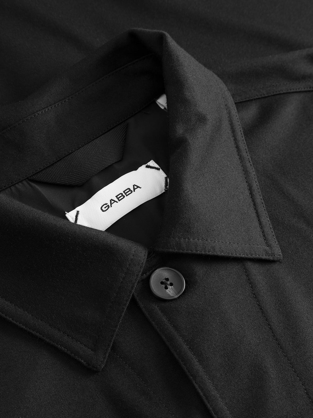 A close-up of the Clipper A Nygo - Black overshirt by Gabba, featuring a black collar, visible button, and an inner white label with the Gabba logo—ideal for everyday wear or a modern layered style.