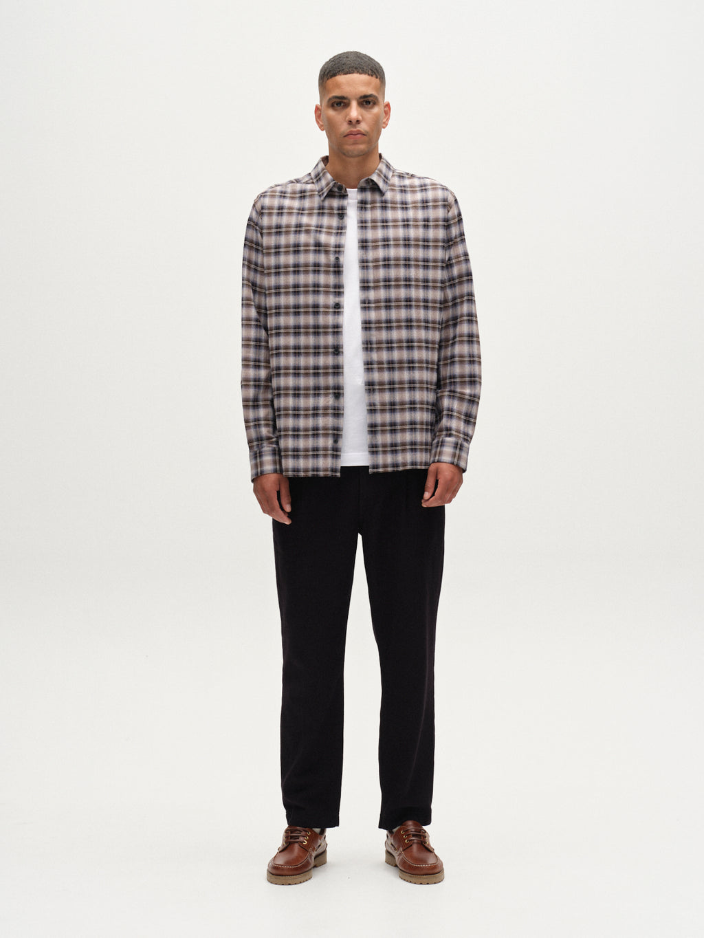 A man stands facing forward against a white background, wearing the Gabba Sven Reg Crepe LS - Blue Check shirt over a white t-shirt, styled with black pants and brown shoes for a smart casual look.
