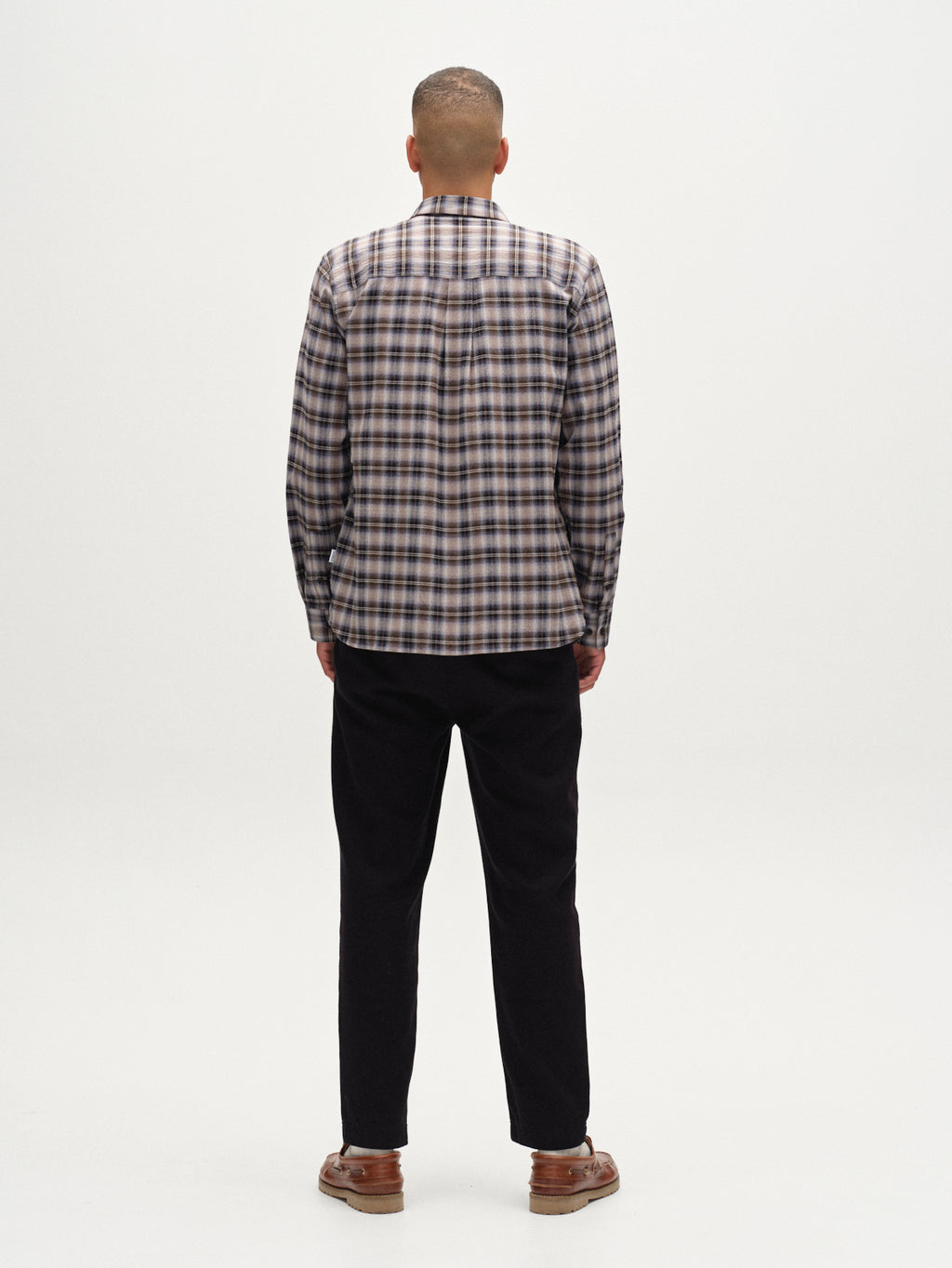 A man faces away from the camera wearing the Gabba Sven Reg Crepe LS - Blue Check shirt, paired with dark pants and brown shoes for a smart casual look against a plain white background.