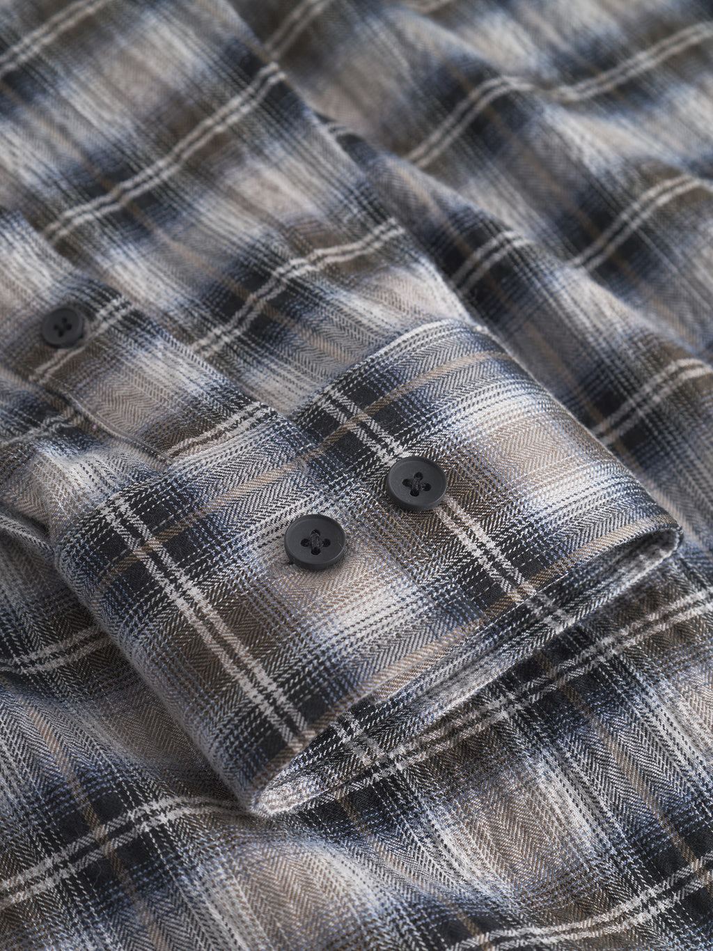 A close-up of the Gabba Sven Reg Crepe LS - Blue Check shirt, featuring folded plaid flannel in blue, gray, and beige tones with a cuff showing two black buttons and fine stitching for a smart casual style.