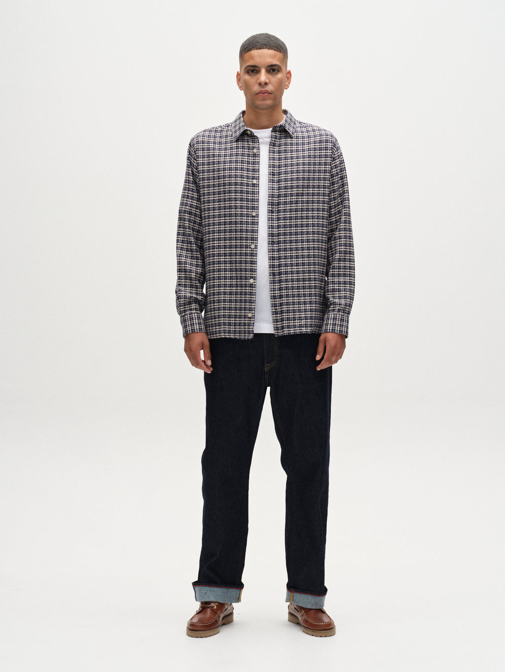 A man faces forward against a white background, wearing the Gabba Sven reg TX LS - Blue Check plaid long-sleeve shirt over a white T-shirt, paired with dark jeans and brown shoes.