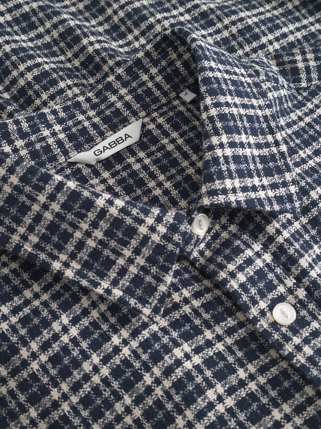 Close-up of the Gabba Sven reg TX LS - Blue Check shirt featuring navy and white checks, white buttons, a collar, and a branded tag at the neckline. This long-sleeve shirt is made from 100% cotton for comfort and style.