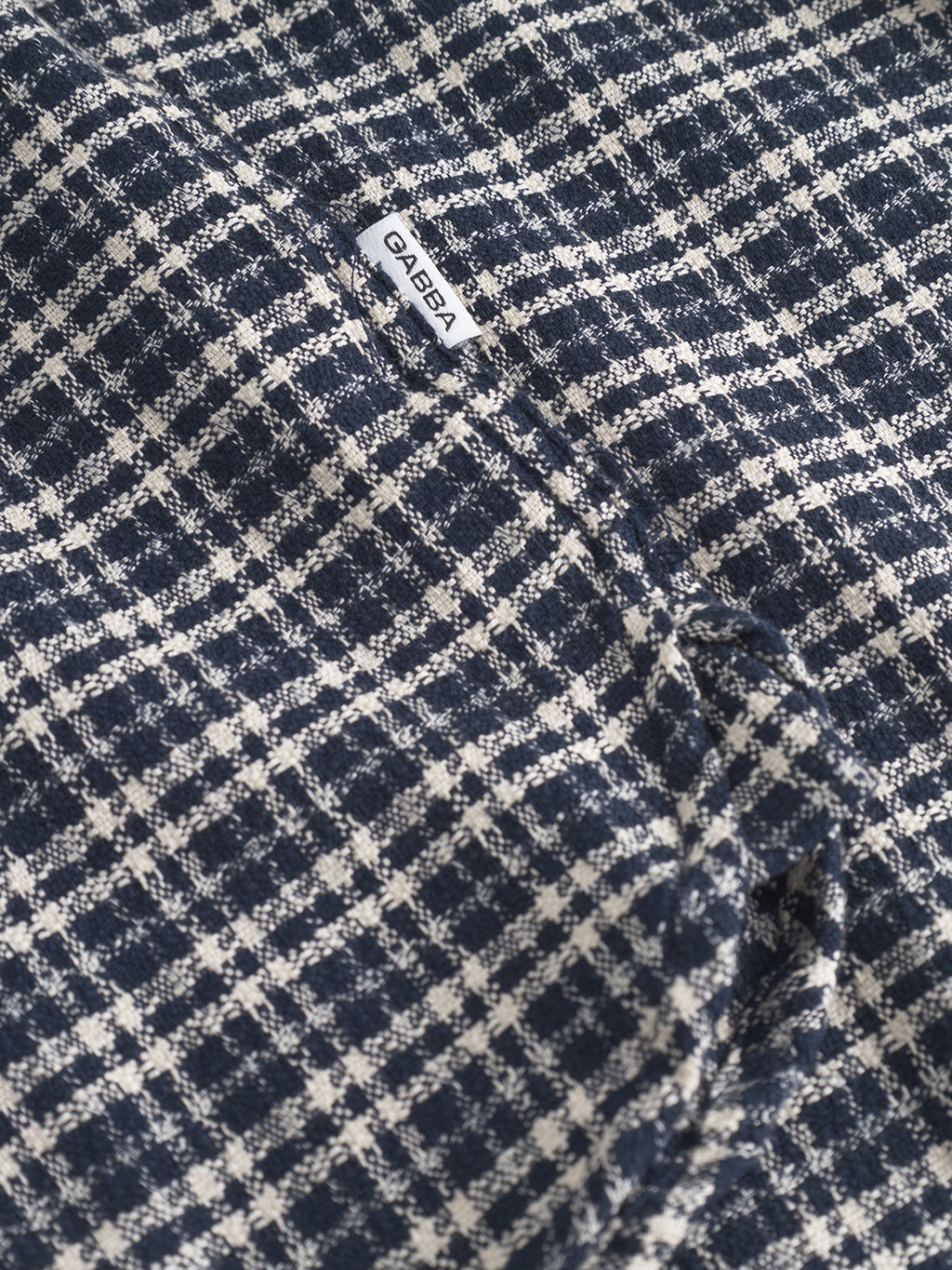 Close-up of the blue and white checkered Sven reg TX LS - Blue Check shirt by Gabba, made from 100% cotton with a small white Gabba label. The texture and woven pattern of this long-sleeve shirt are clearly shown.