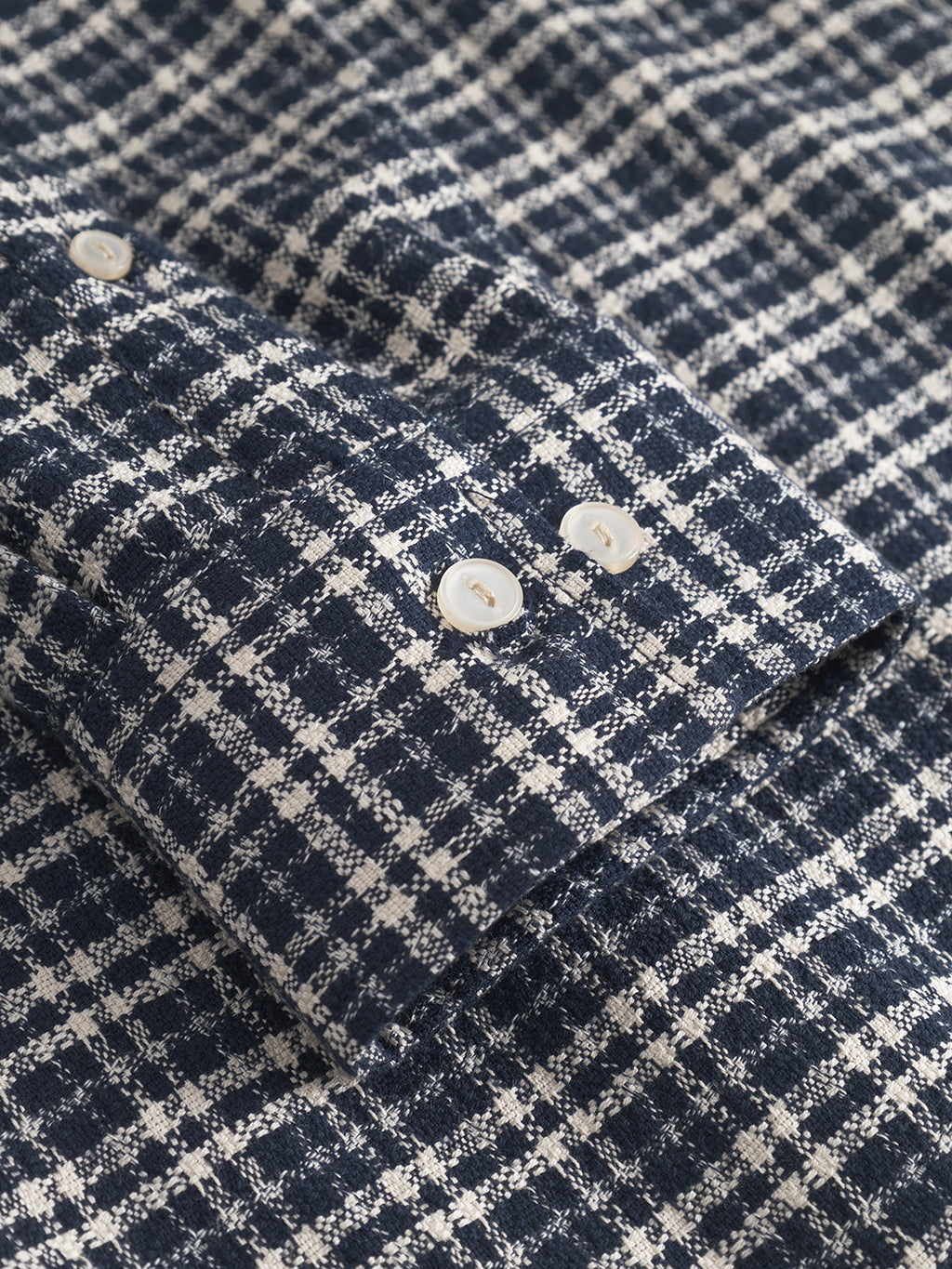 Close-up of the Gabba Sven reg TX LS - Blue Check shirt, featuring a textured navy and white check pattern, 100% cotton fabric, long sleeves, and a cuff finished with two white buttons.