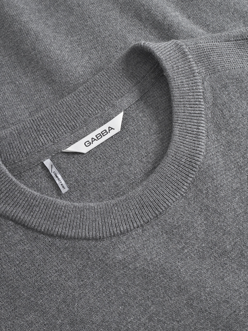 A close-up of the Gabba Blanko New LS sweater in dark grey melange shows its soft Milano knit fabric, ribbed crew neck, GABBA neck label, and a visible side tag. The texture appears smooth and refined.