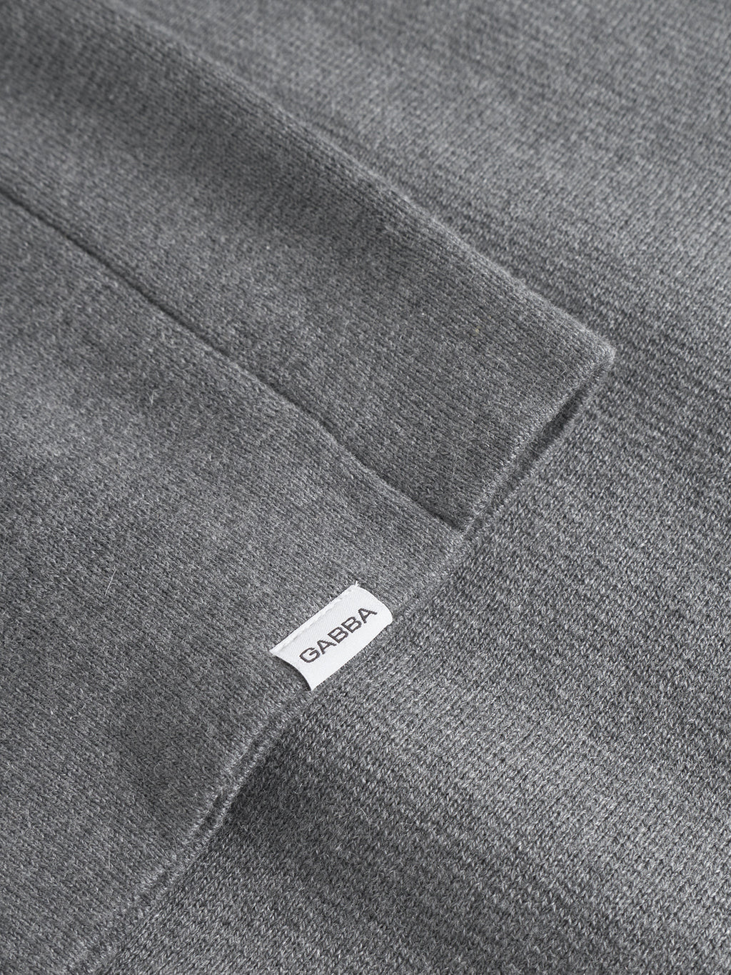 Close-up of the Blanko New LS - Dk. Grey Mel by Gabba shows Milano knit fabric in dark grey with a visible seam and a small white “GABBA” tag sewn onto the knitwear.