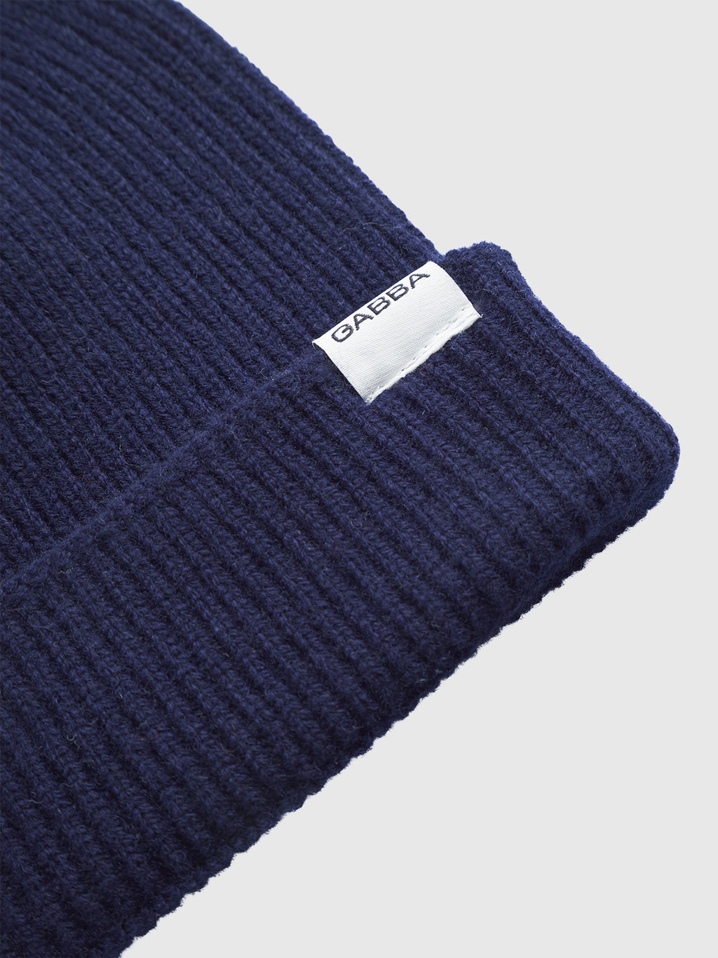 Close-up of the Biella Aico - Navy beanie by Gabba, crafted from soft merino wool in a ribbed knit with folded brim and a white Gabba tag on the edge. This cozy cold-weather essential is set against a light gray background.