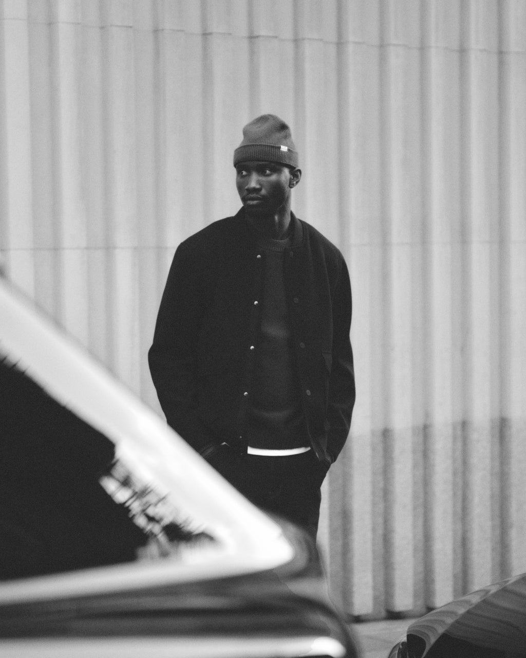 A man in a Gabba Biella Aico - Green ribbed wool hat and dark jacket stands on a city street, looking to the side. The black and white photo has a blurred car in the foreground.