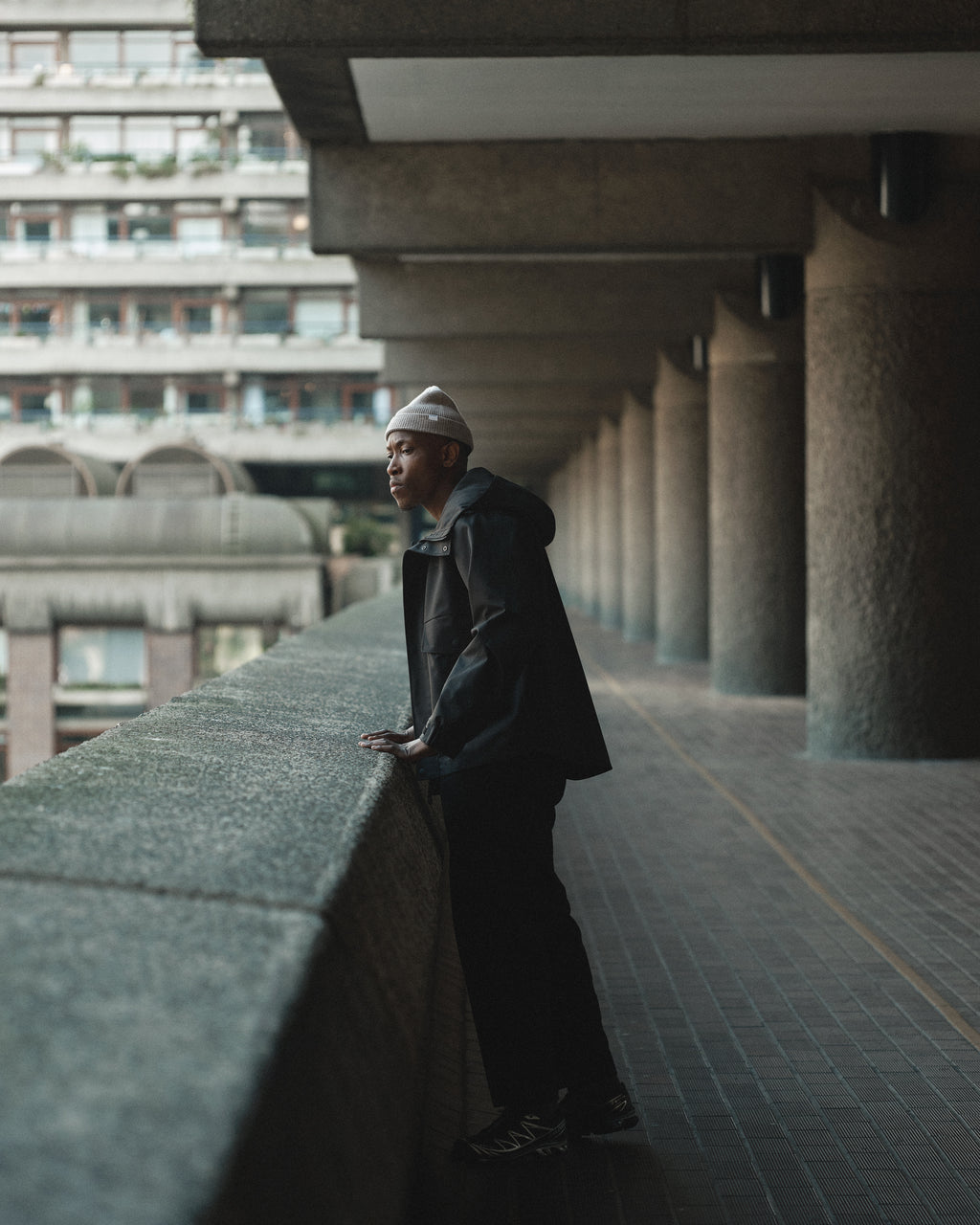 Wearing the Gabba Biella Aico Lt. Brown beanie and a dark jacket, a person leans on a concrete railing in an urban setting with modern buildings and columns in the background.