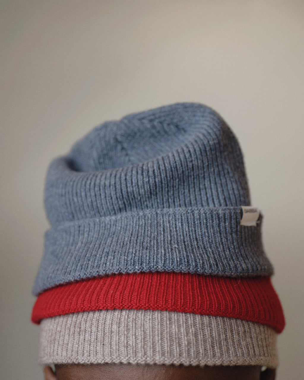 A close-up shows a person wearing three stacked knit beanies, with the Gabba Biella Aico - Vintage Indigo beanie featuring a small white tag on top. The background is softly blurred.