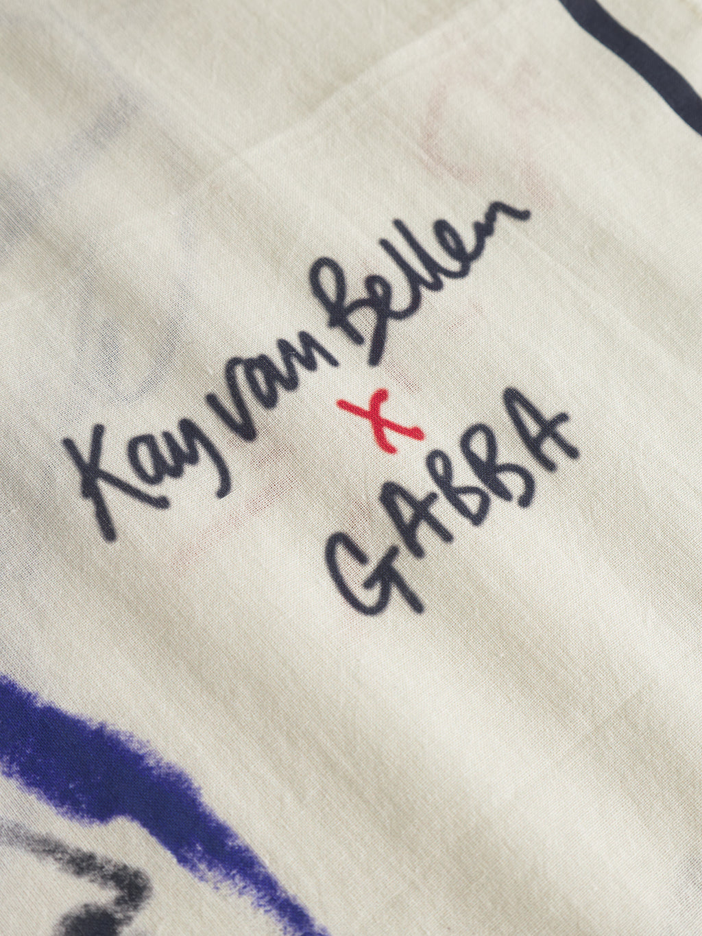A close-up of the Bandana Kay - Multi Pattern by Gabba, showing white fabric with handwritten black text Kay van Bellen x GABBA, where the x is written in red.