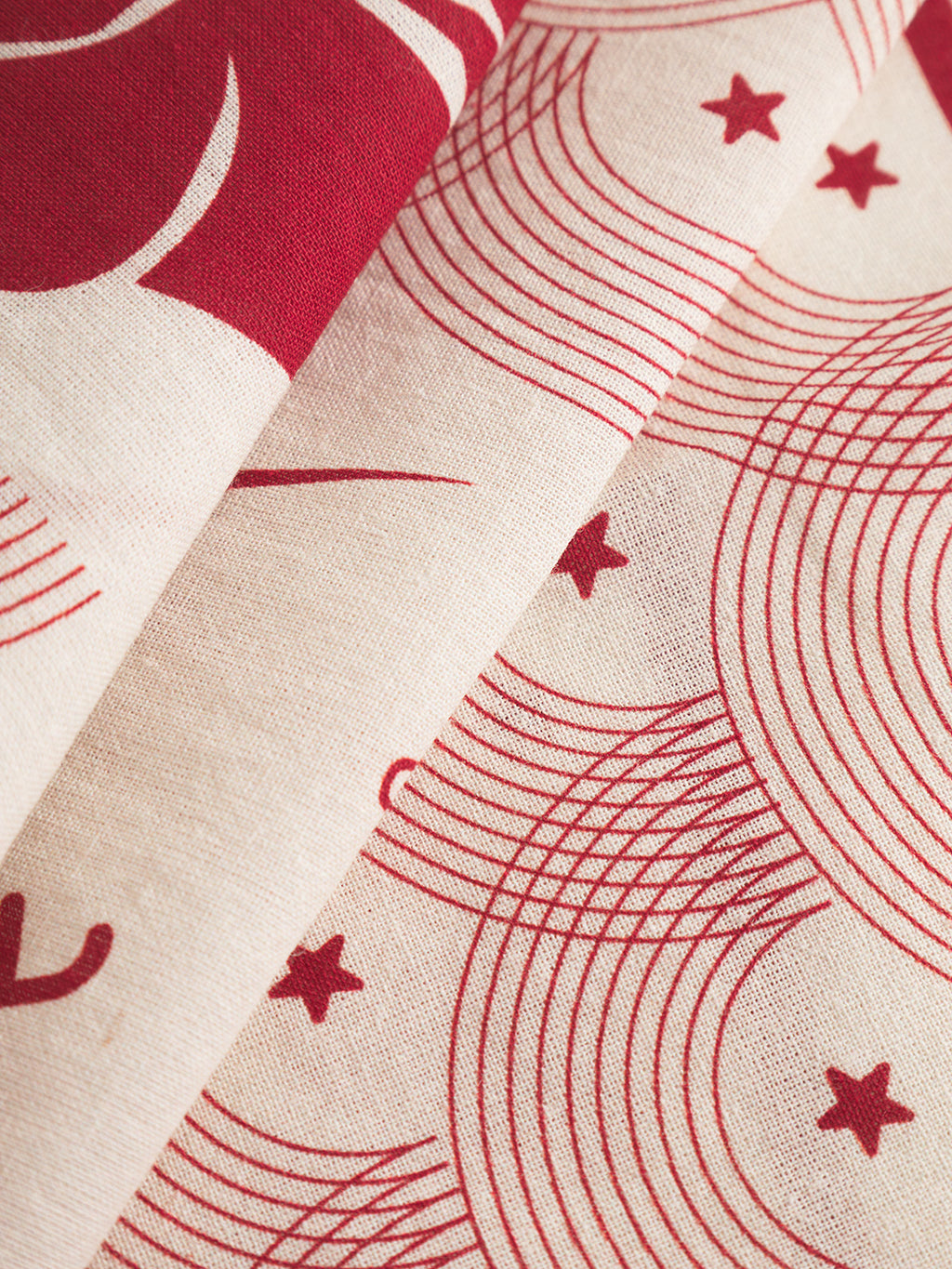 A close-up of Gabba’s Bandana Waves - Multi Pattern fabric shows beige material with red, abstract curved lines and small stars. The Japanese-inspired print and folded layers evoke the look of a classic cotton bandana.