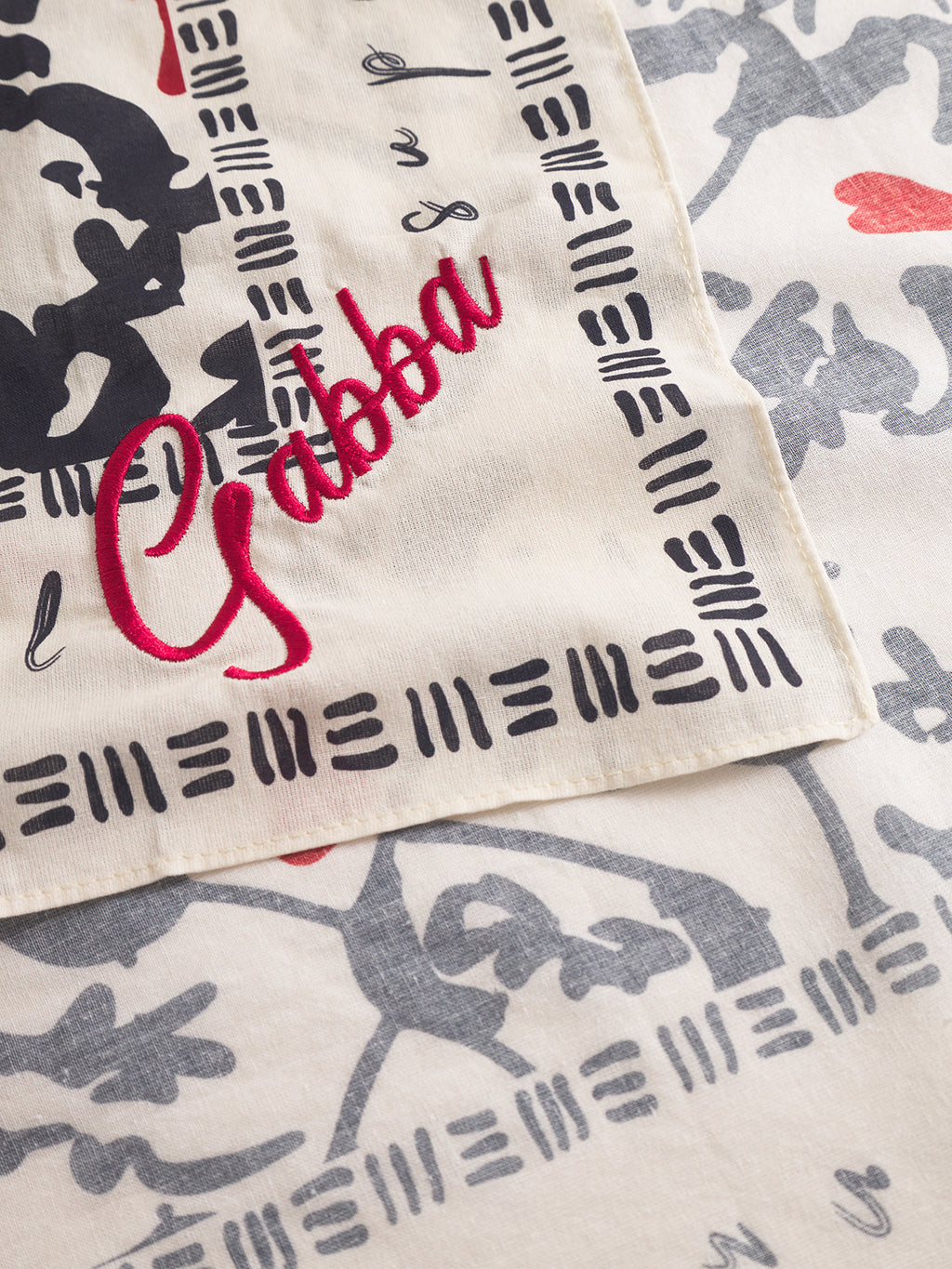 A close-up of the Gabba Bandana Yovi - Multi Pattern shows premium cotton fabric with black geometric and Japanese-inspired prints, plus “Gabba” in red script embroidery on a corner. Another patterned fabric is partially visible beneath it.