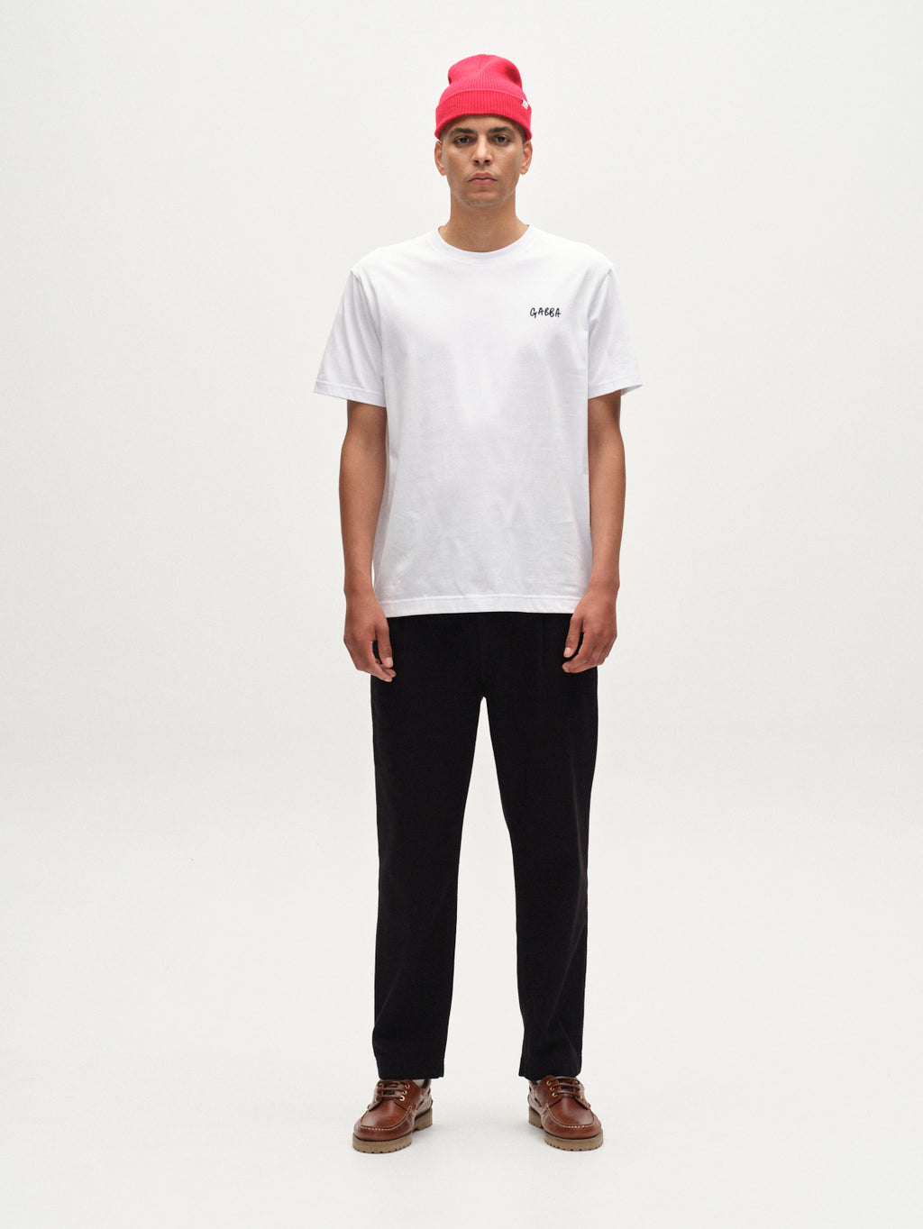 A person stands facing forward against a plain light background, wearing a bright pink beanie, black pants, brown shoes, and the Gabba Dune Kay Dot SS GOTS T-shirt in white with small black text.