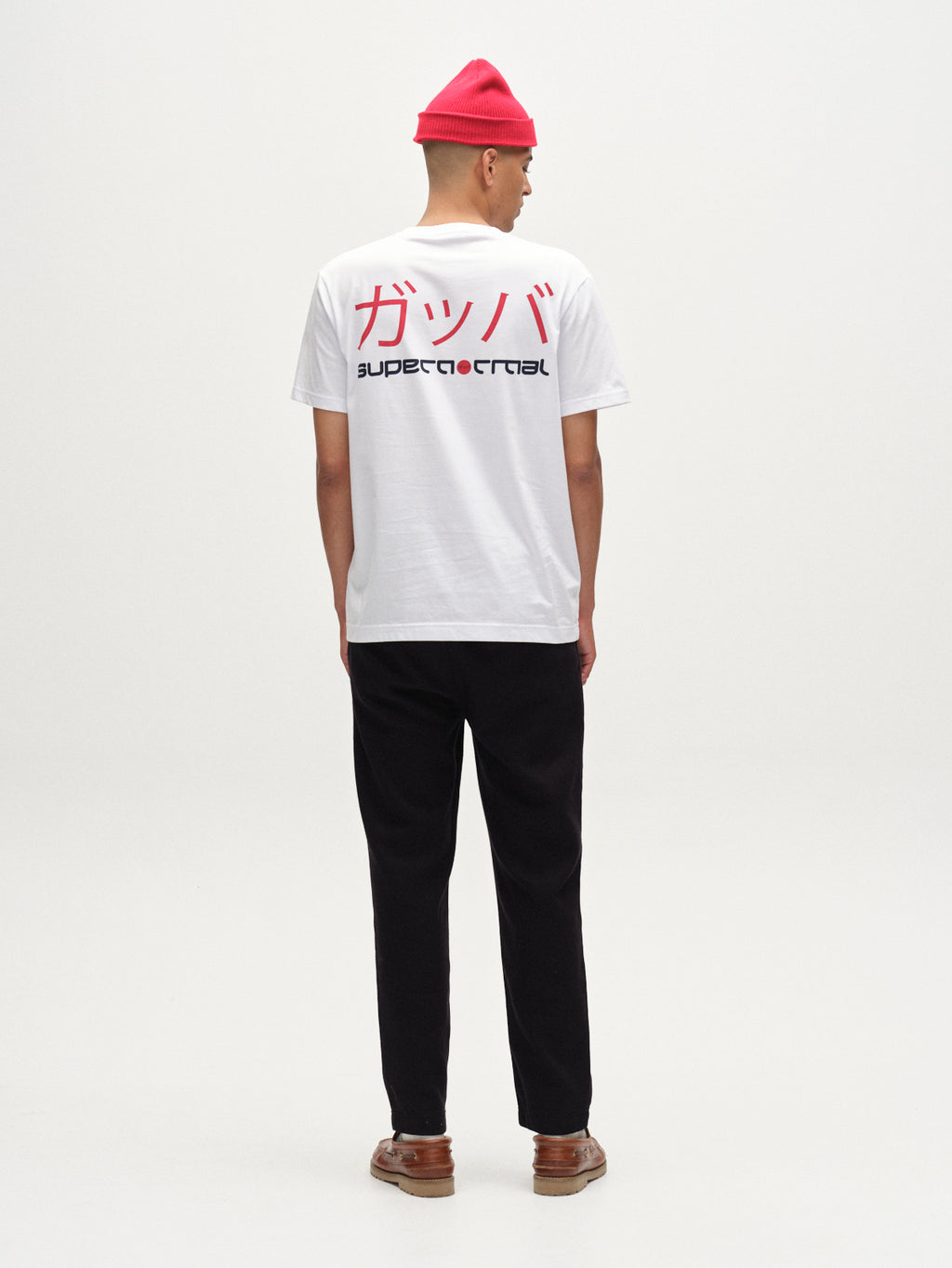 A person stands facing away in a Gabba Dune Kay Dot SS GOTS - White T-shirt with red Japanese and black SUPERCRITICAL text on the back, black pants, brown shoes, and a bright pink beanie against a plain white background.