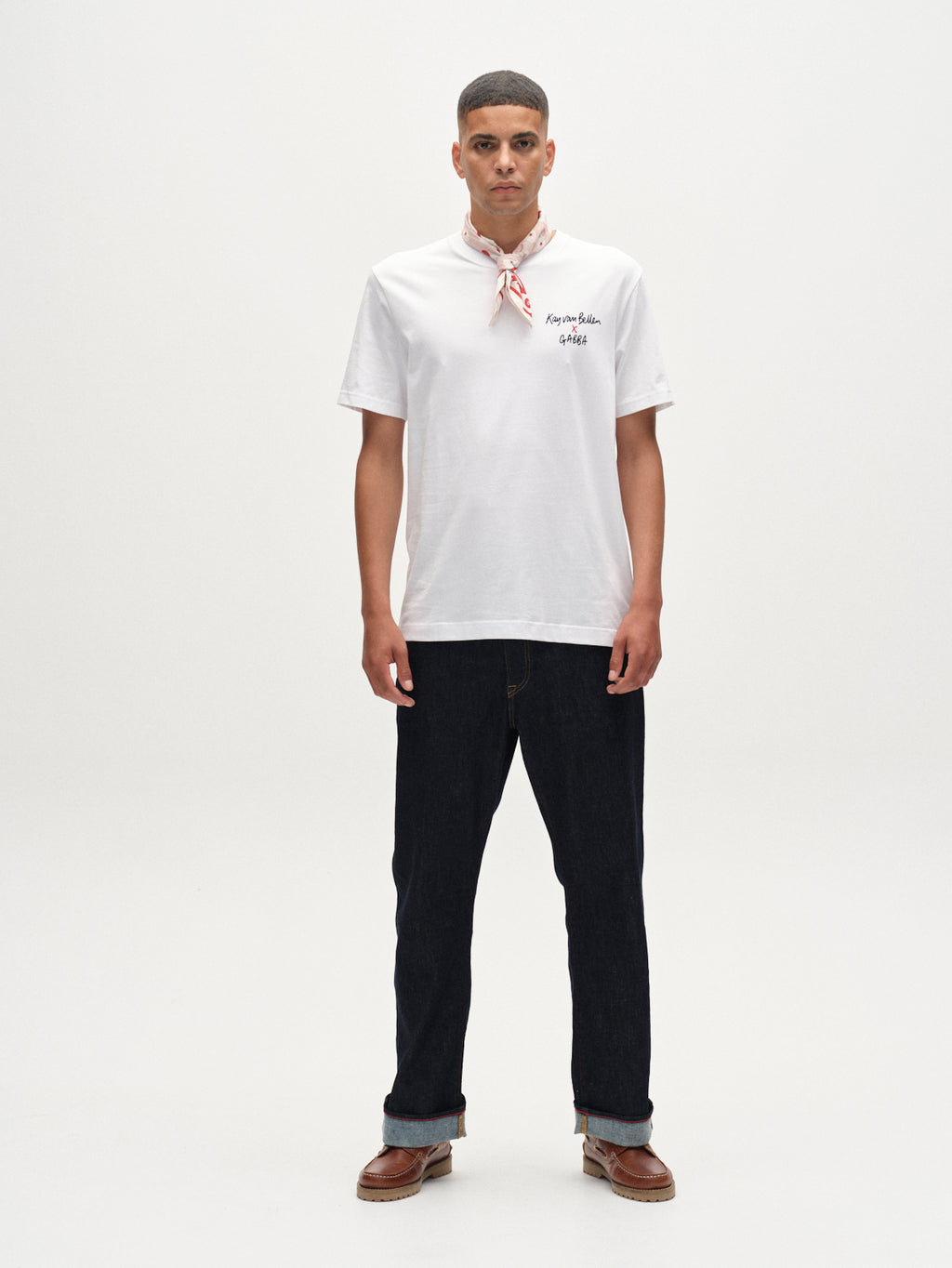 A young man stands facing forward against a plain white background, wearing the Gabba Dune Kay Man SS GOTS tee in white with text on the left chest, a red-patterned neck scarf, dark jeans, and brown shoes.