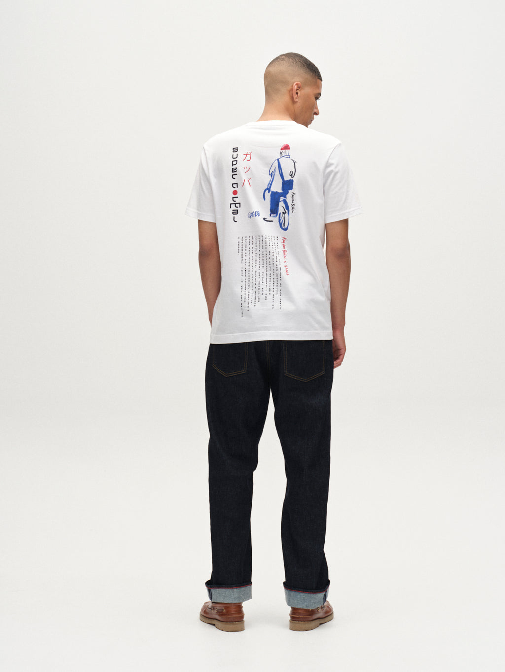 A person stands facing away, wearing a Gabba Dune Kay Man SS GOTS tee in white with blue and red graphic art and Japanese text on the back, paired with dark jeans and brown shoes, against a plain white background.
