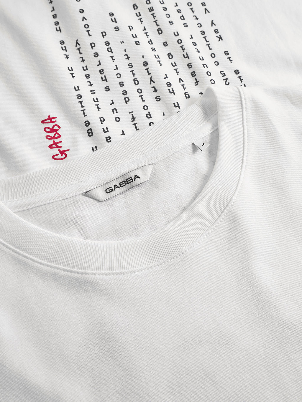 Close-up of the white Gabba Dune Kay Man SS GOTS tee, featuring the brand label on the collar and a vertical black chest print with Gabba in red among black letters. Made from GOTS-certified fabric for comfort and style.