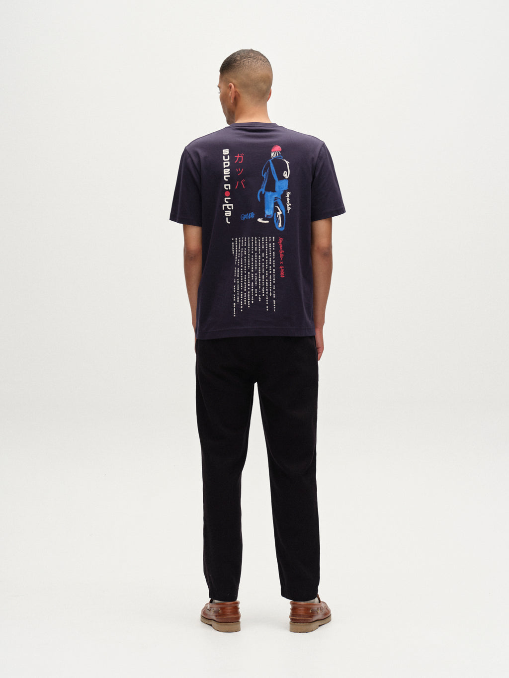 A person faces away, wearing the Gabba Dune Kay Man SS GOTS navy organic cotton t-shirt with colorful Japanese-inspired minimalist graphics on the back, paired with black pants and brown shoes against a plain white background.