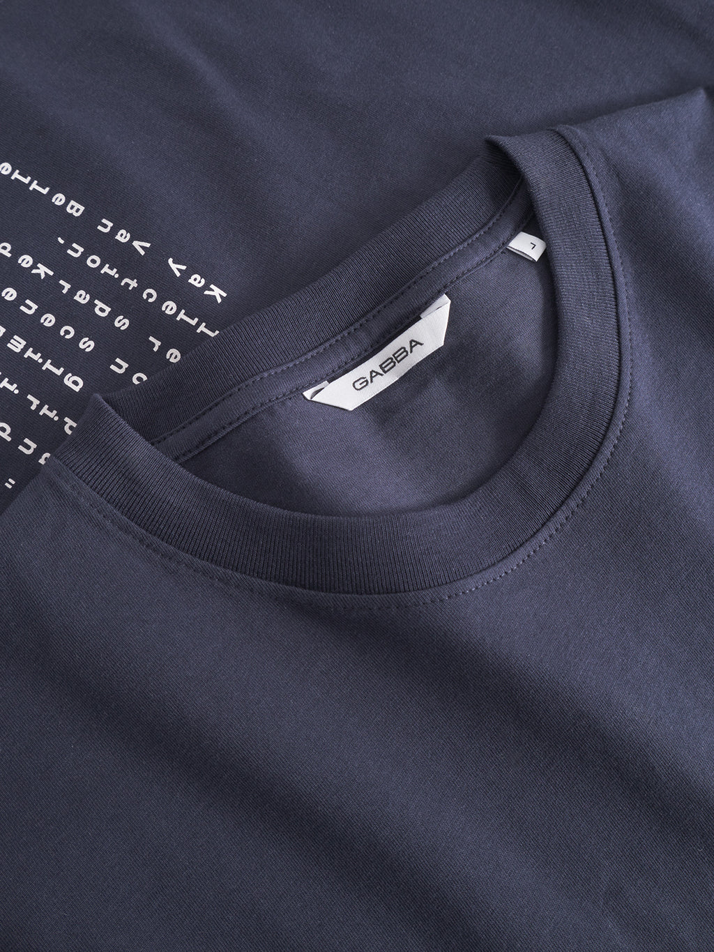 A close-up of two Gabba Dune Kay Man SS GOTS - Navy t-shirts stacked, one with a white GASBA label and the other partially revealing a column of white symbols—echoing Japanese-inspired minimalism.