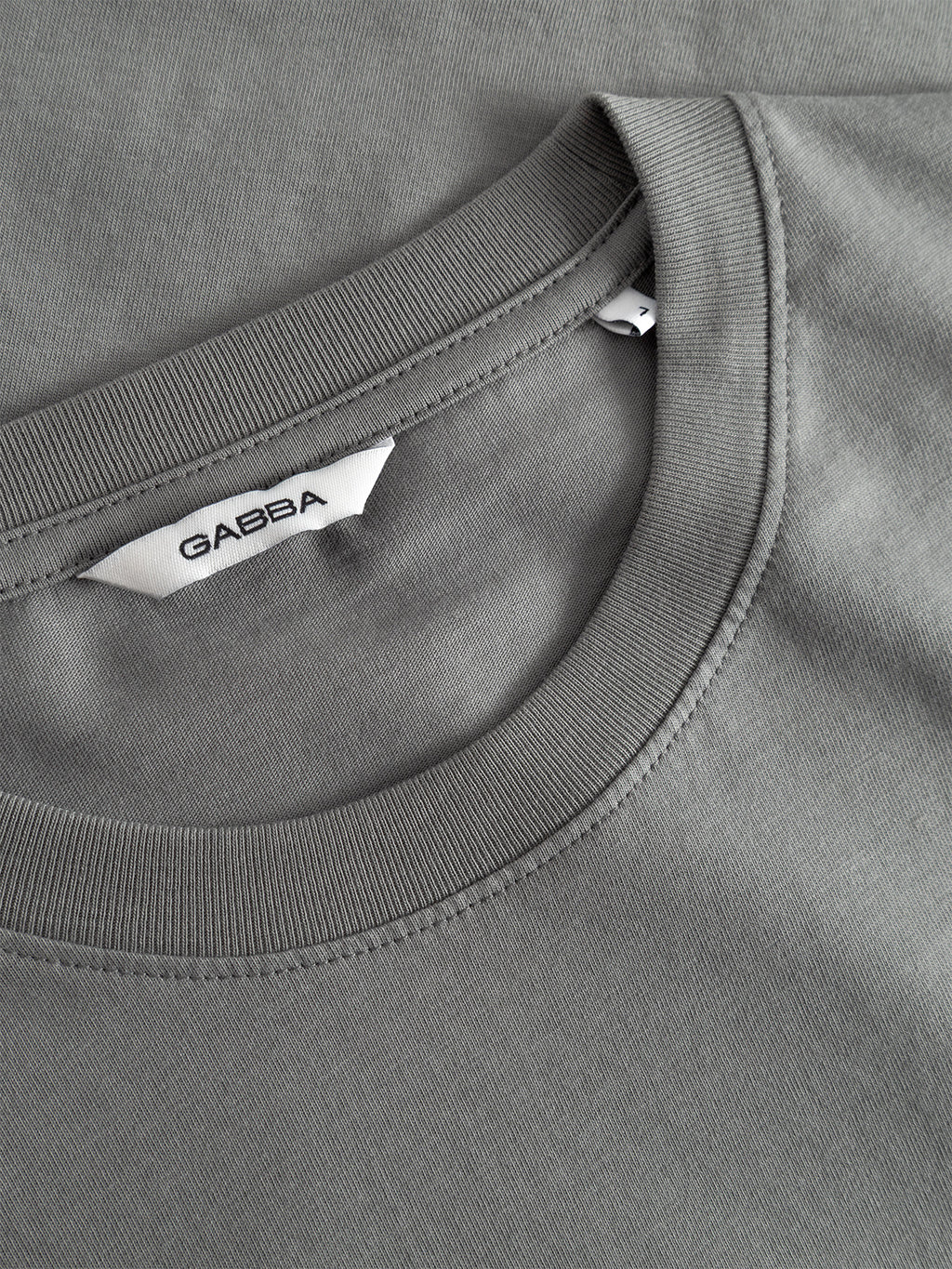 Close-up of a Dust Sage Green crew neck organic tee, the Dune Kay Man SS GOTS by Gabba, with visible stitching and a white collar label showing the brand name Gabba in black letters.