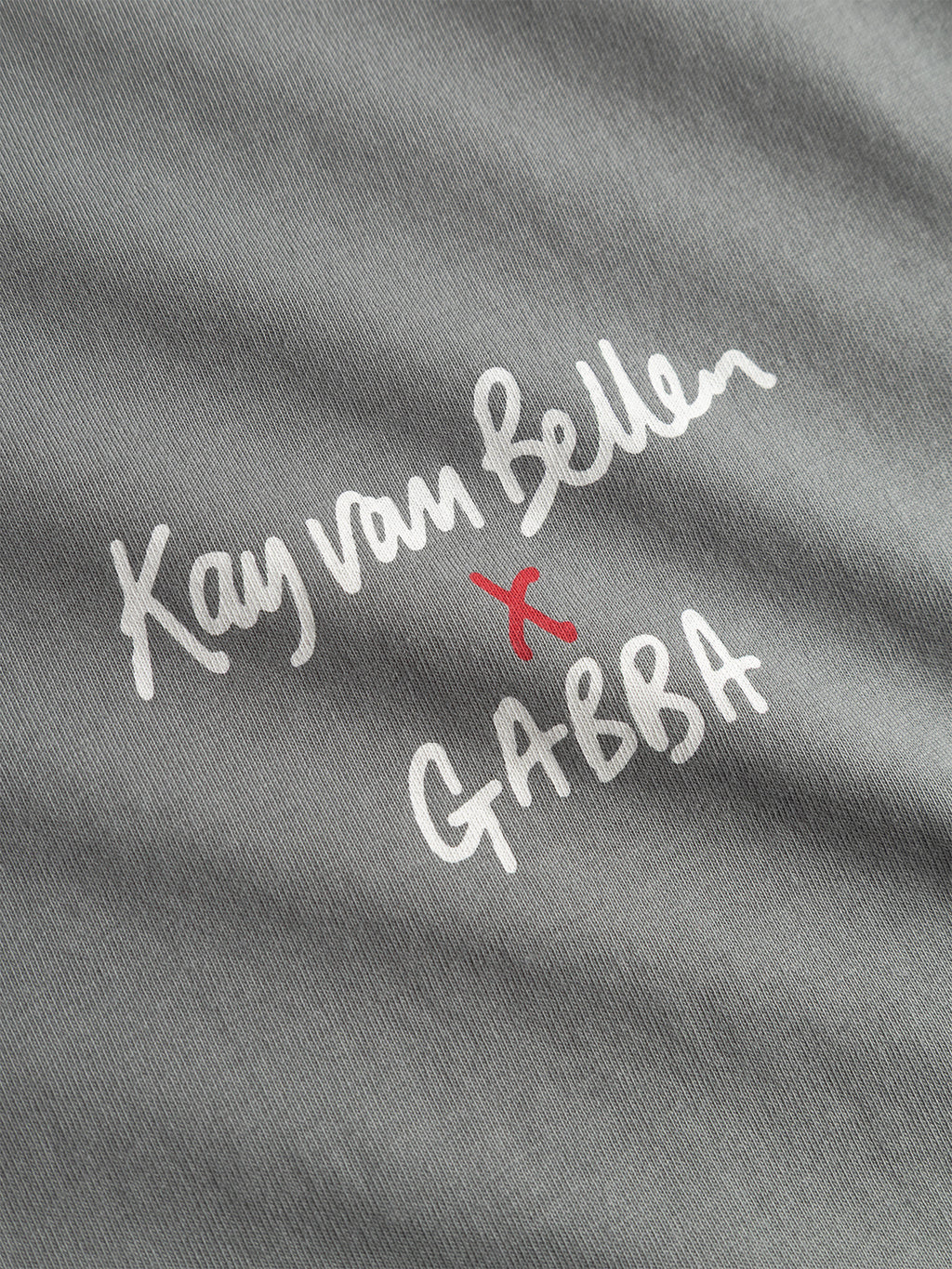 Dust sage green organic tee by Gabba, GOTS-certified cotton, with white handwritten Kay van Bellen x GABBA text and a red x marking the artist collaboration. Product: Dune Kay Man SS GOTS.