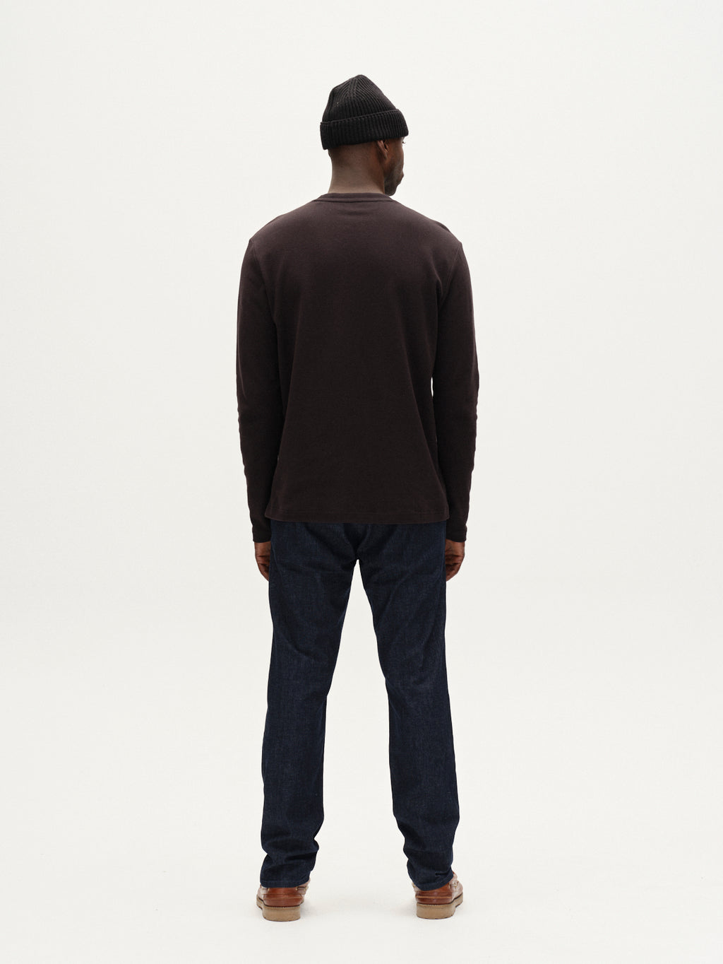 A man stands in a minimal, white studio wearing Gabba’s Paul Sisu - Dark Denim slim fit jeans, paired with a black beanie, dark long-sleeve shirt, and brown shoes. He faces away from the camera.