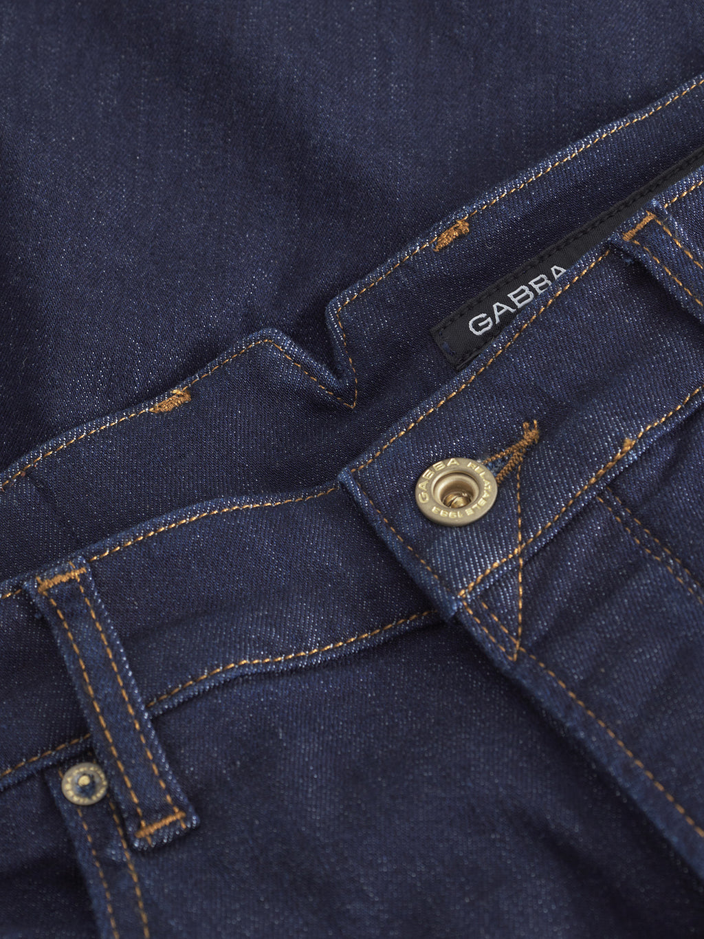 Close-up of blue tapered denim jeans, featuring visible orange stitching, a metal button, and a black label with the Gabba brand—Alex Just - Denim Blue—crafted from recycled fibers.