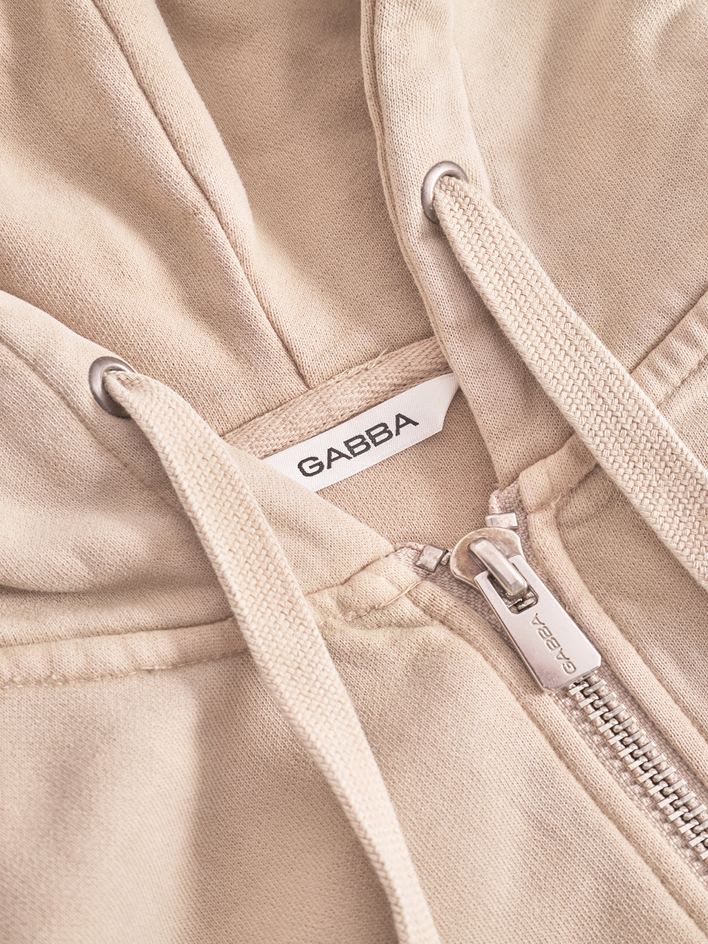 A close-up of the Delphi Bump GWC hoodie in sand by Gabba, featuring a metal zipper, drawstrings, and a white Gabba label inside the collar—ideal for effortless casual layering.