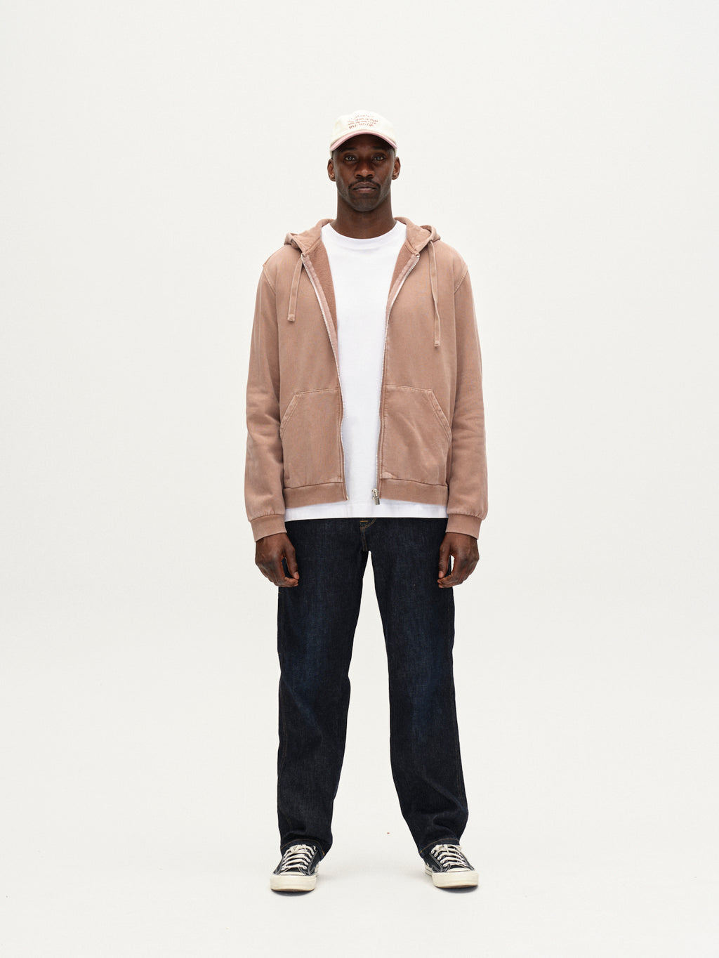 A man faces forward in a modern casual look, wearing the Gabba Delphi Bump GWC - Rose hoodie over a white T-shirt, dark jeans, black and white sneakers, and a light baseball cap against a plain white background.