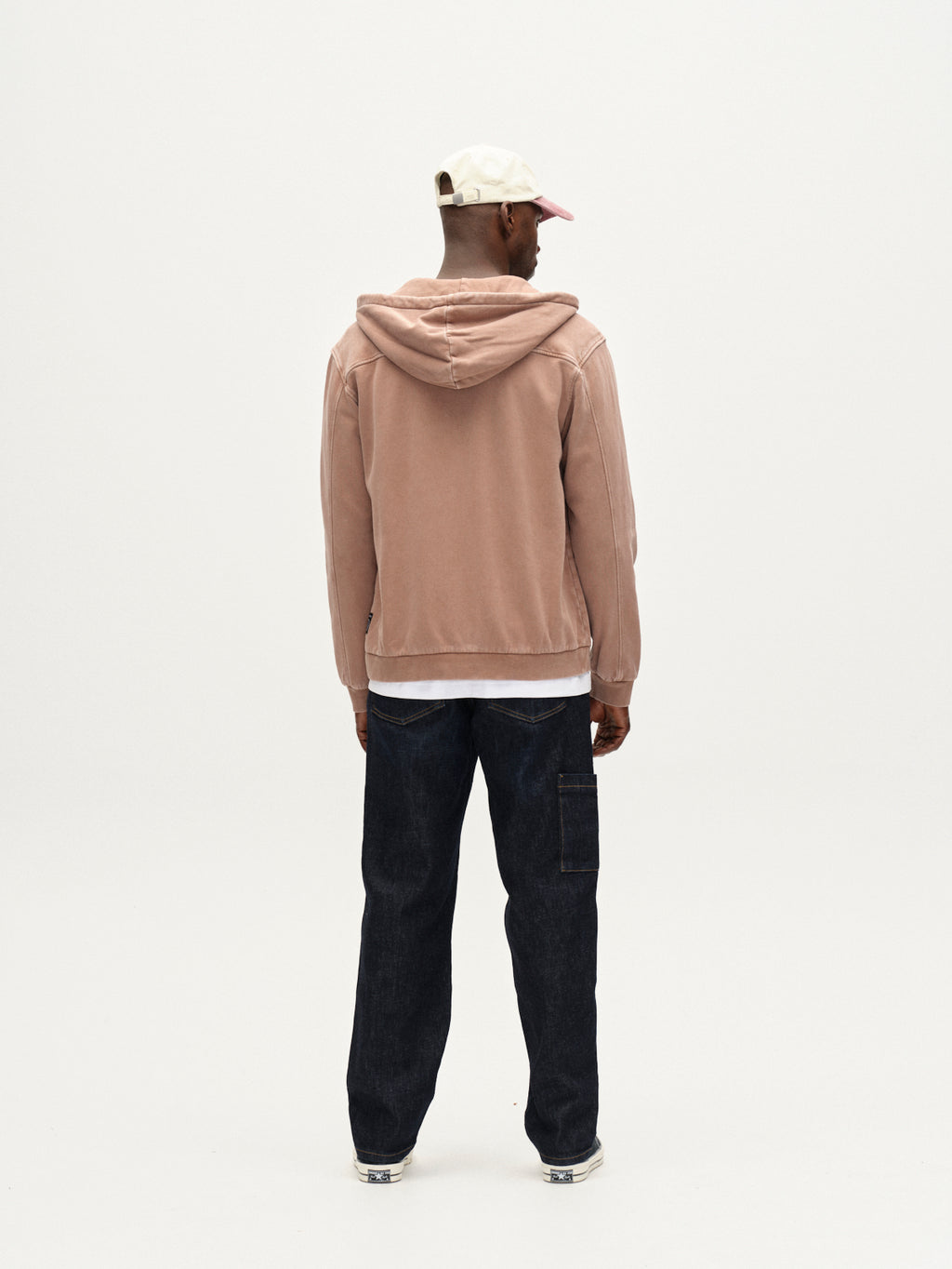 A person faces away from the camera in a modern casual look, wearing a light pink Delphi Bump GWC - Rose hoodie from Gabba, dark jeans, a white shirt underneath, and a white and pink baseball cap against a plain white background.