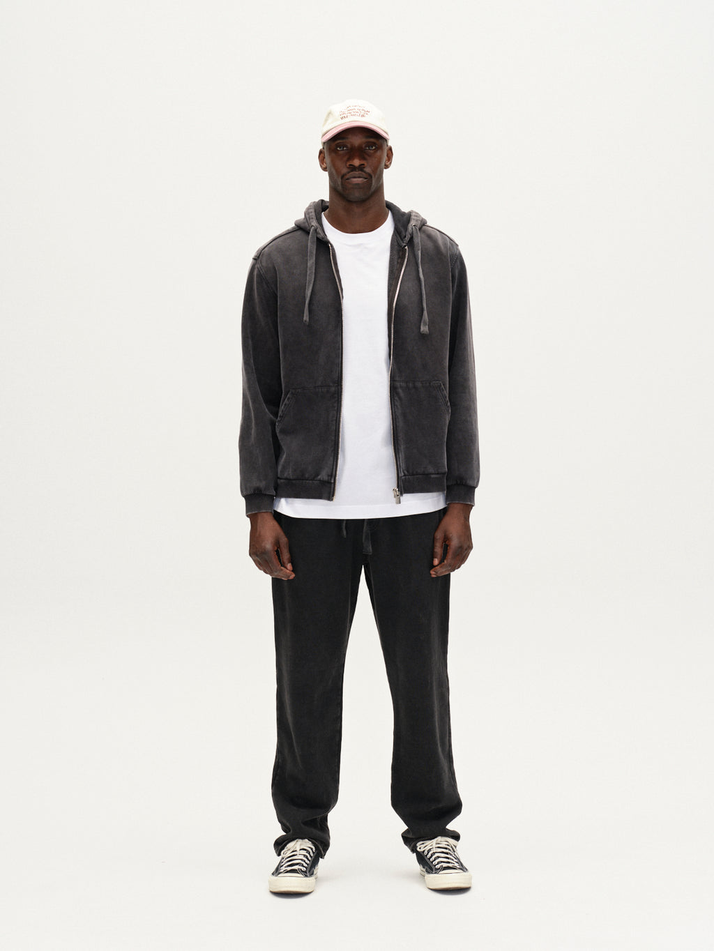 A man faces forward against a plain white background, wearing Gabbas Delphi Bump GWC - Black zip hoodie, a white t-shirt, black pants, black and white sneakers, and a light-colored baseball cap.