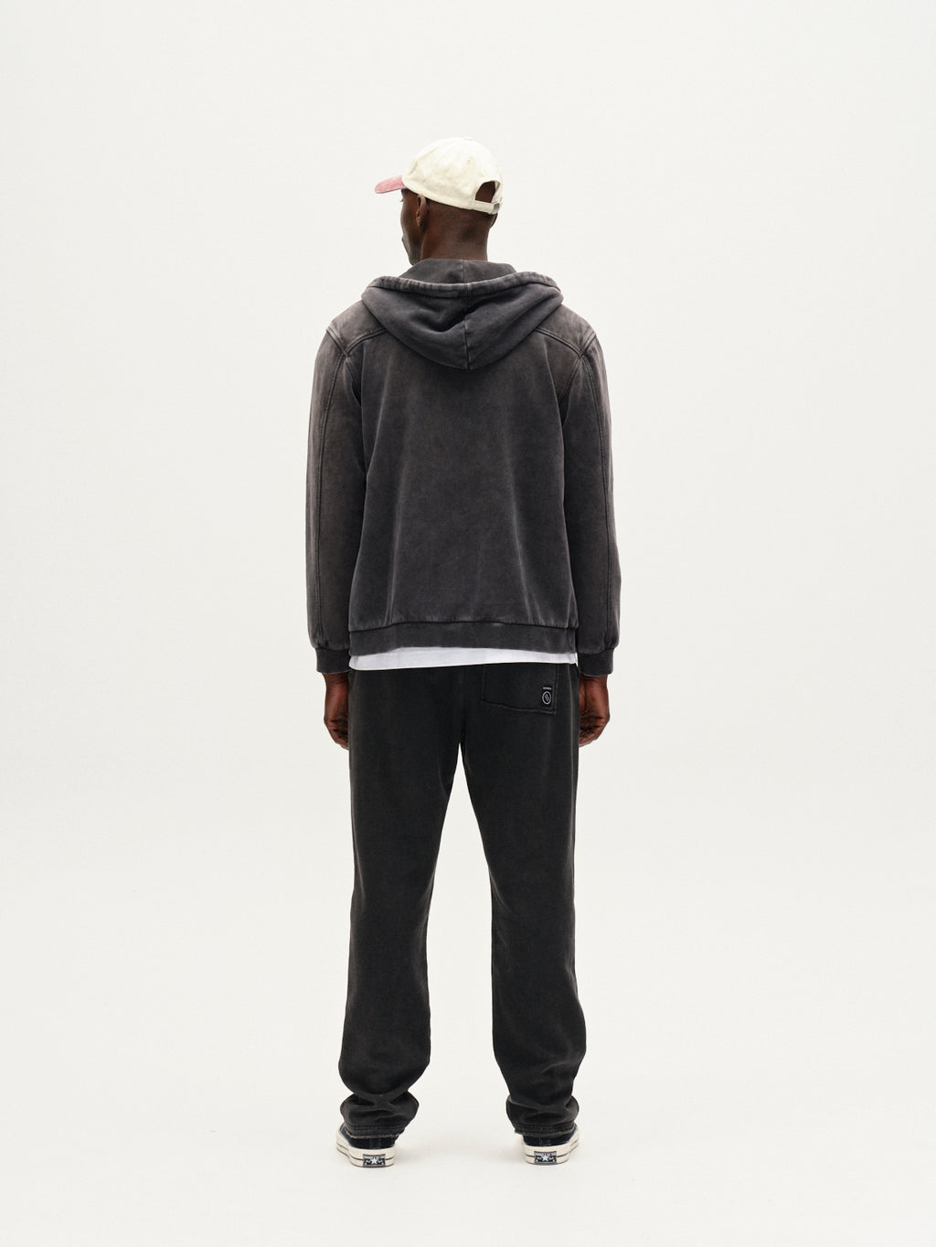 A person stands facing away from the camera wearing Gabba’s Delphi Bump GWC - Black hoodie, dark sweatpants, a white cap, and sneakers against a plain white background.