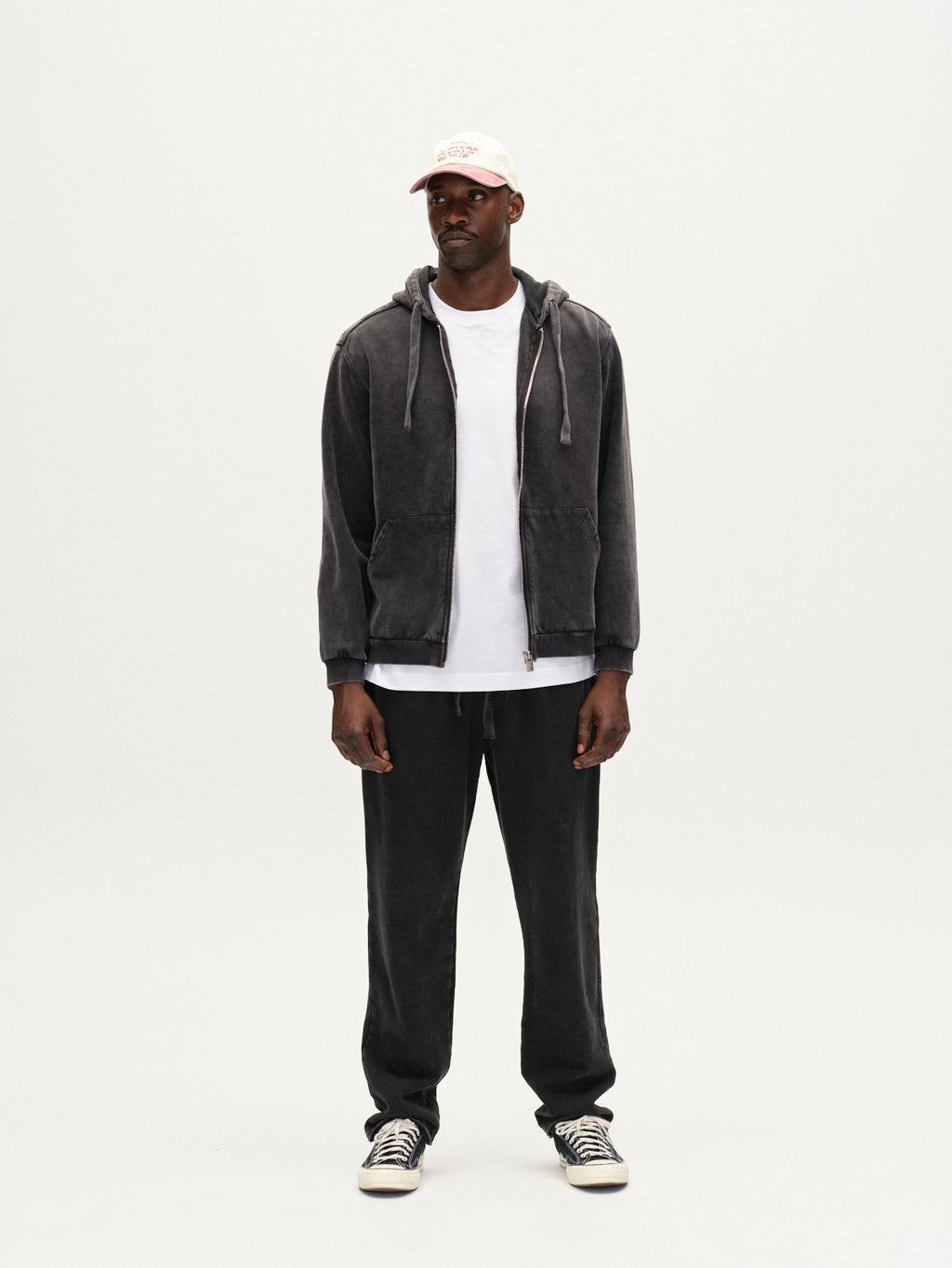 A man stands against a white background wearing a white cap, dark gray zip-up hoodie, white t-shirt, loose straight-fit pants with an elastic waistband, and Gabba Pireo Bump GWC - Black sneakers. He looks straight ahead with a neutral expression.