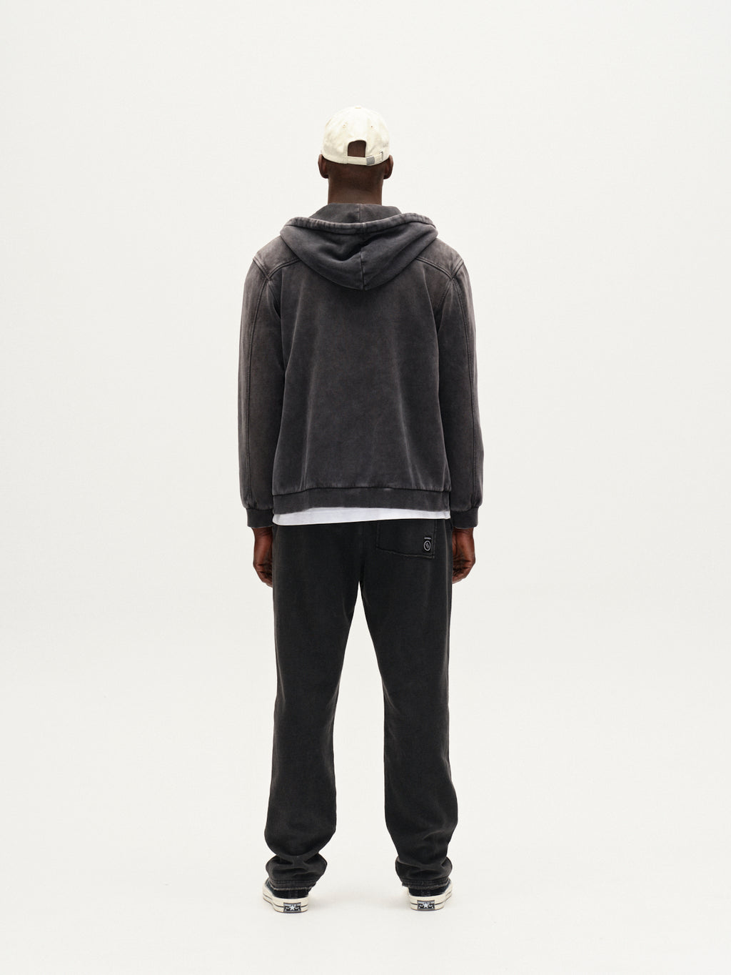A person stands facing away against a white background, wearing Gabbas Pireo Bump GWC - Black hoodie, loose straight-fit pants with an elastic waistband, a white shirt, white cap, and sneakers.