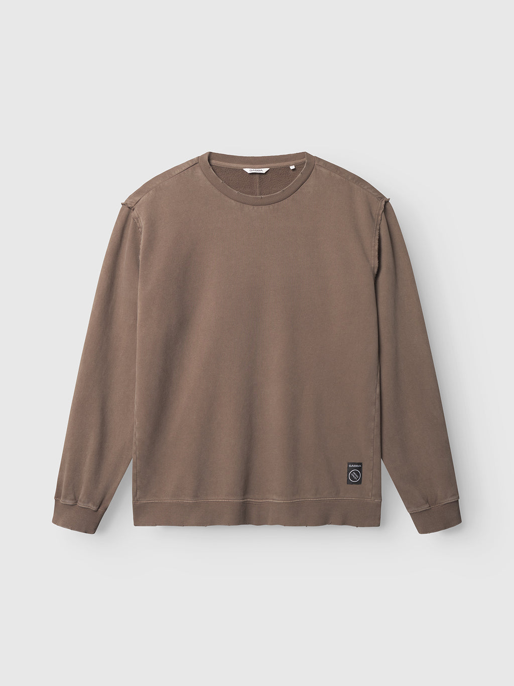 The Gabba Volos Bump GWC - Brown is a super-soft, long-sleeve crew neck sweatshirt with visible seams, ribbed cuffs and collar, and a small black patch near the lower left hem, shown laid flat on a light background.
