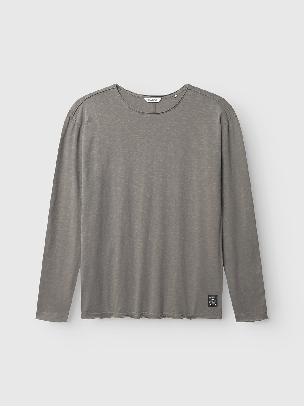 The Gabba Paros Rif GWC - Grey is a super soft cotton long sleeve t-shirt with a round neckline, laid flat on a white background. It features a small rectangular patch with a circular design near the bottom hem.