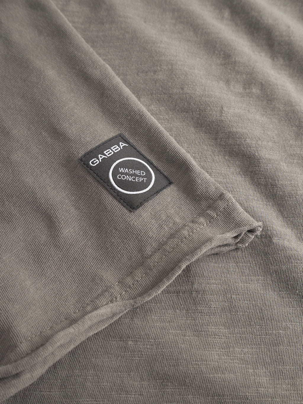 Close-up of Paros Rif GWC - Grey by Gabba: gray textured fabric with a black “GABBA WASHED CONCEPT” patch, on a super soft, naturally faded long sleeve t-shirt.