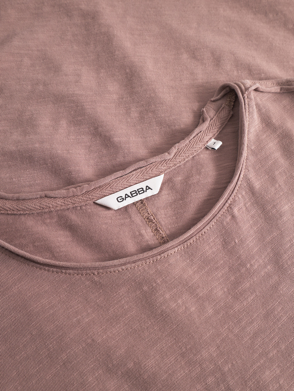 Close-up of the Gabba Paros Rif GWC - Rose long sleeve t-shirt in mauve, featuring a round neckline and a white label with black Gabba text sewn inside the collar. The super soft cotton fabric looks slightly textured and comfortable.