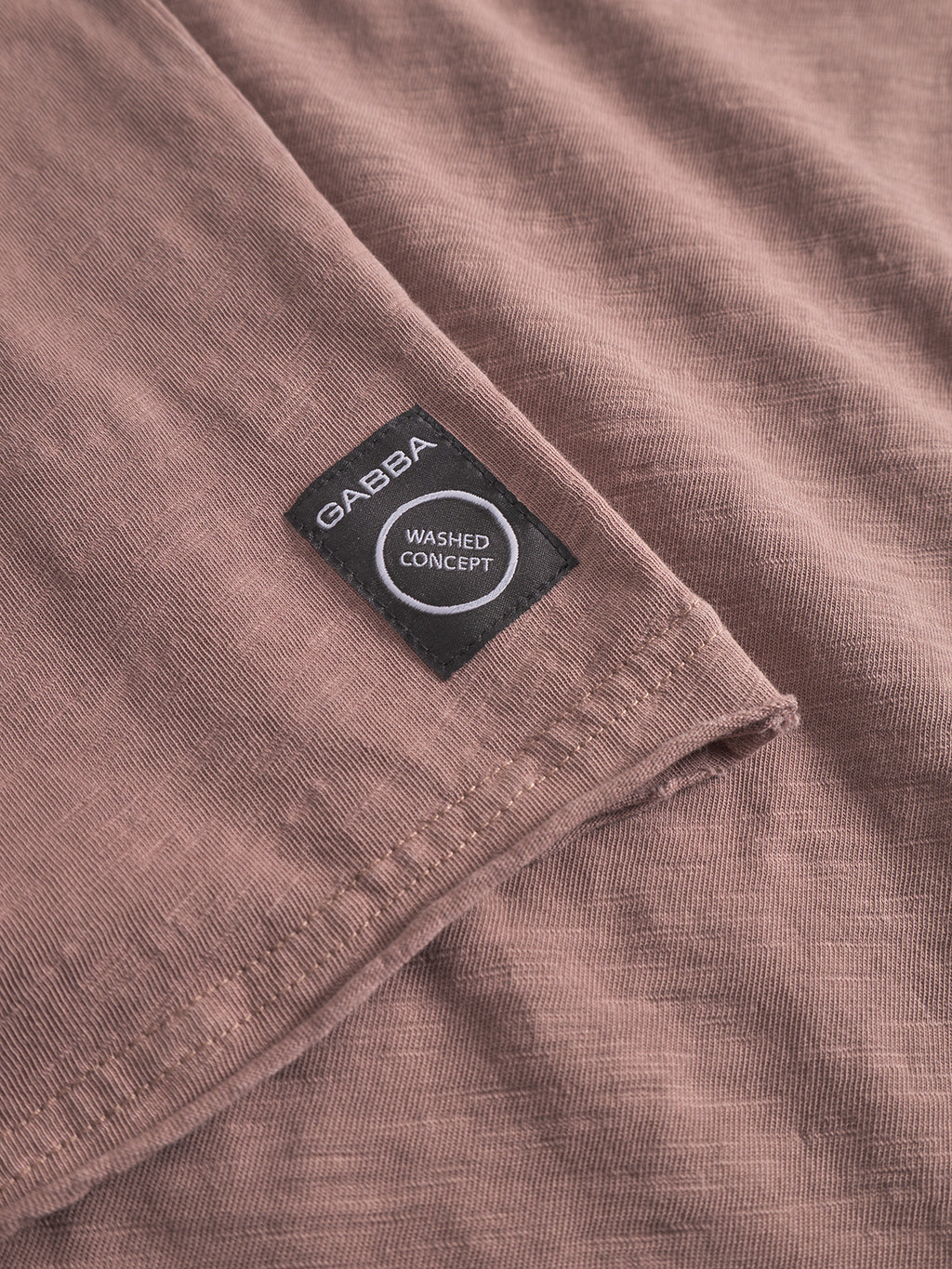Close-up of the Paros Rif GWC - Rose by Gabba, showing its textured mauve cotton fabric and a black rectangular Gabba Washed Concept label stitched near the seam, hinting at a faded look and long-sleeve t-shirt style.