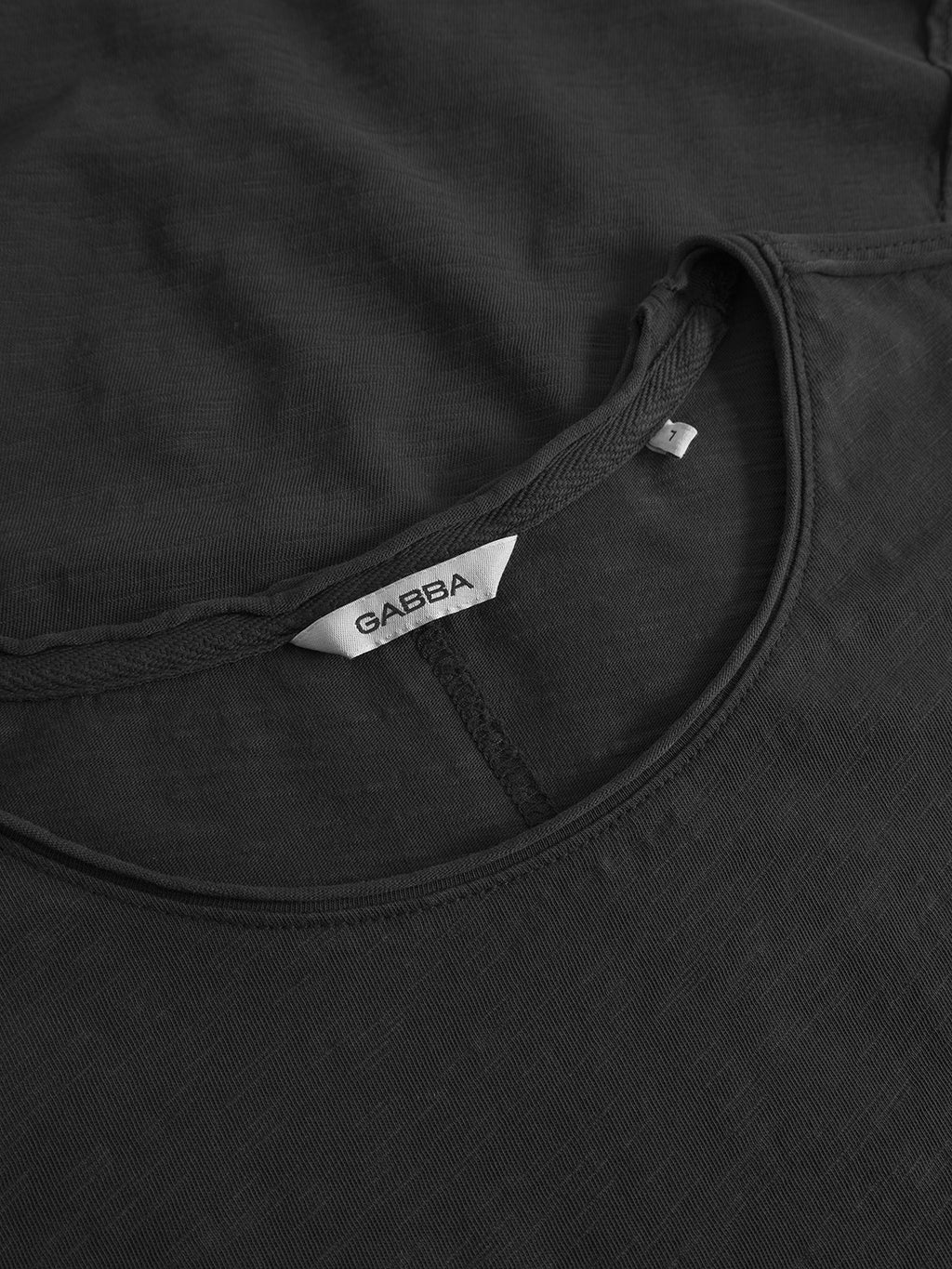 Close-up of the Gabba Paros Rif GWC - Black cotton t-shirt, featuring a dark gray faded look. The shirt is laid flat, highlighting the neckline, visible stitching, and a white inner label marked GABBA.