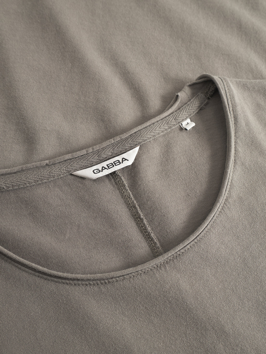 Close-up of the Gabba Idra Rig GWC - Grey t-shirt laid flat, highlighting its collar and stitching. Crafted from super soft cotton, it has a white Gabba label and an L size tag inside the neckline.