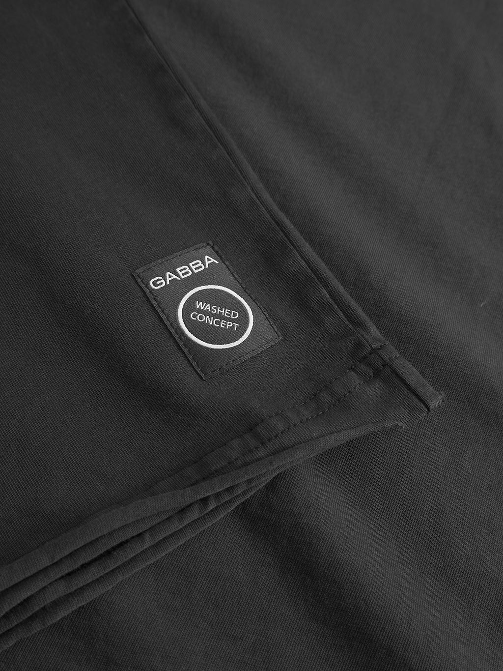 Close-up of folded black Idra Rig GWC tee by Gabba, showing a rectangular black label with white “Gabba” and “Washed Concept” text, stitched on super soft vintage faded cotton.