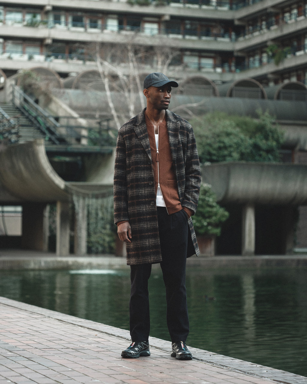 A man wearing a Gabba Sity Self Cap in dark denim, a plaid overcoat, brown jacket, white shirt, black recycled-fiber pants, and black shoes stands by a pond with modern concrete buildings and greenery in the background.