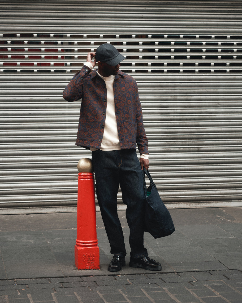 A man in a patterned jacket, white shirt, black cap, and dark blue denim jeans stands by a red post on a city sidewalk, holding the Gabba Bag Self - Dark Blue Denim tote bag, with a metal shutter in the background.