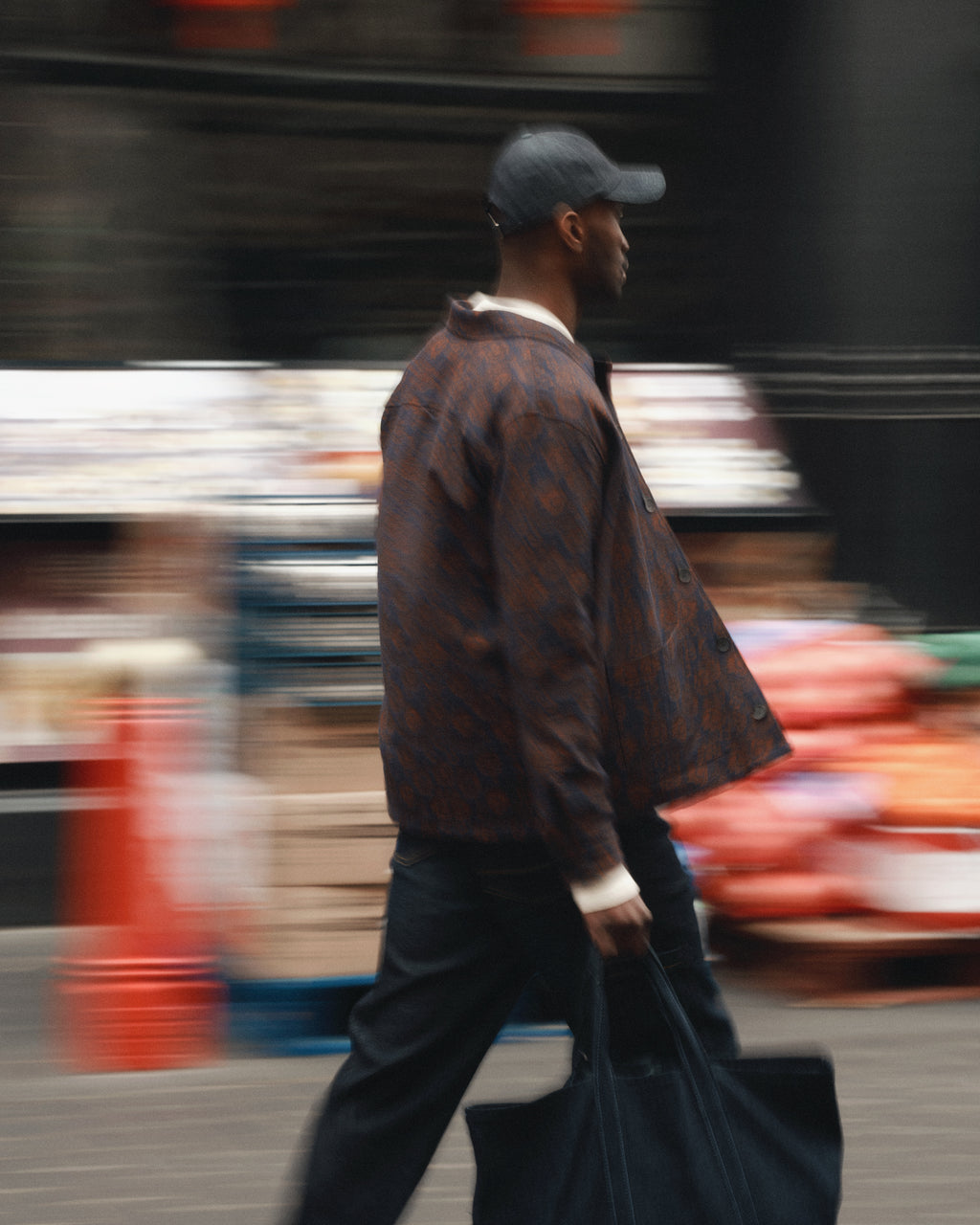 A person wearing a patterned jacket of recycled fibers, black pants, and a black cap walks briskly outdoors in an urban setting, carrying the Gabba Bag Self in dark blue denim. The blurred background suggests movement and city life.