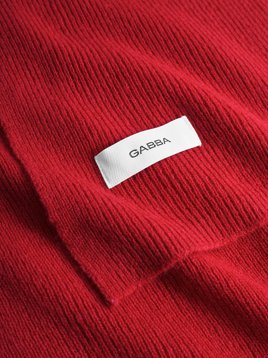 Close-up of Biwa Aico - Red by Gabba: textured red knit scarf fabric with a white rectangular label featuring Gabba in black capital letters.