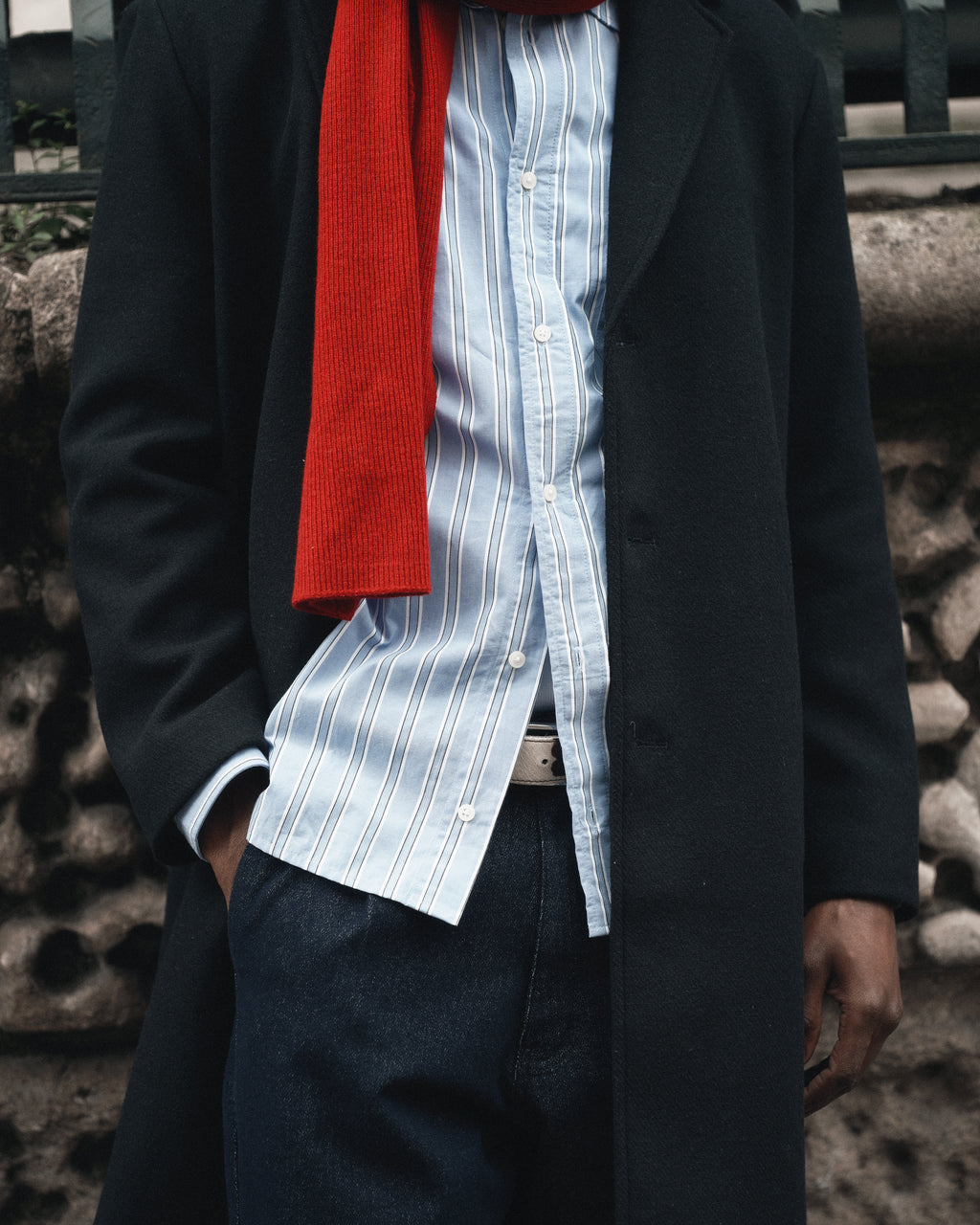 Wearing Gabba’s Biwa Aico - Red scarf with a dark coat, blue and white striped shirt, and dark jeans, a person stands in front of a stone wall with one hand in their pocket; their face is not visible.