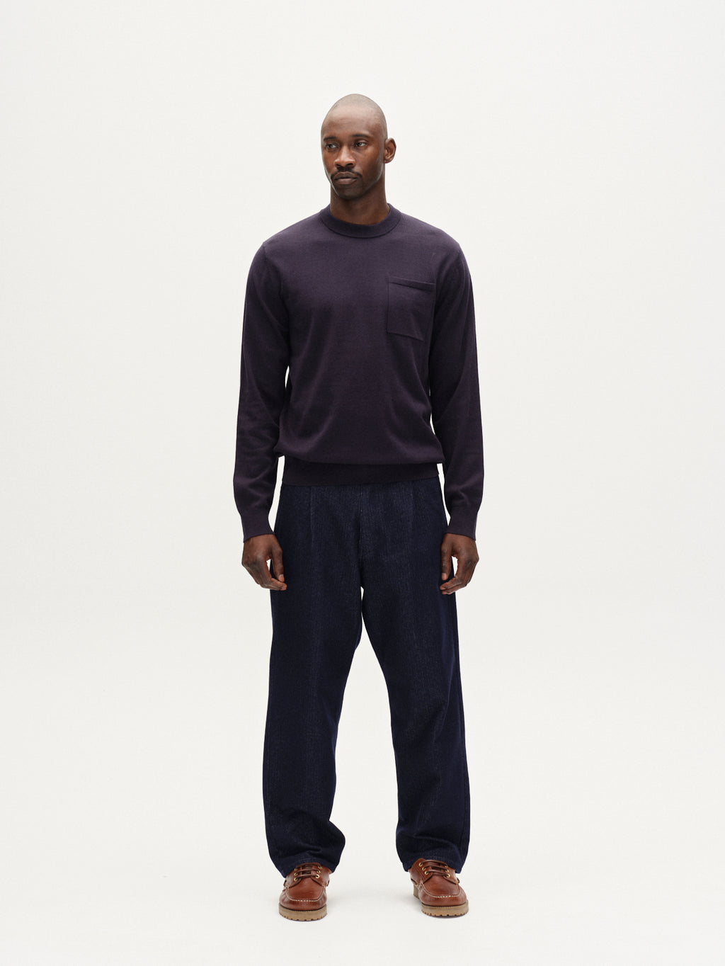 A man stands facing forward against a plain white background, wearing the Gabba Amori Prex LS sweater in navy, loose-fitting dark pants, and brown shoes. He has a neutral expression and his hands rest by his sides.